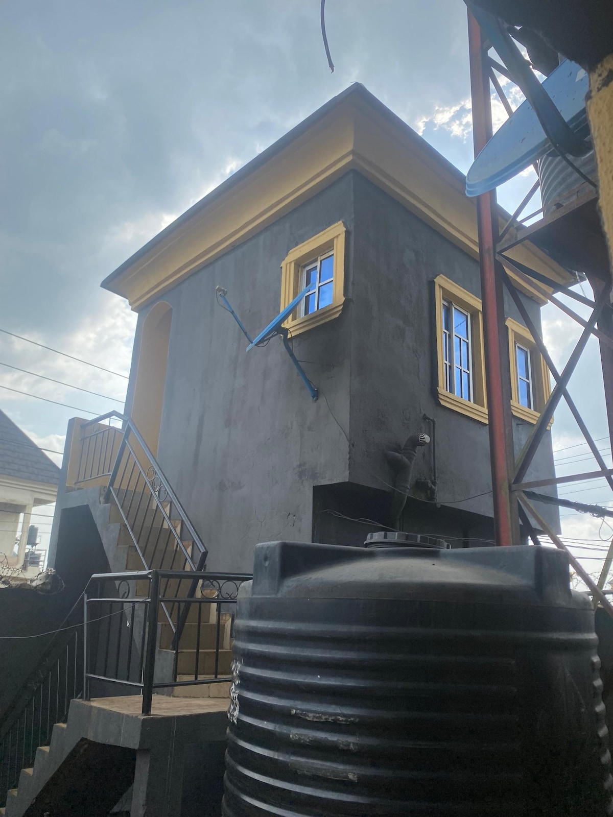 The image features a compact structure with a modern design, showcasing yellow-framed windows that contrast against a dark exterior. A staircase leads to the entrance, while a large water tank is positioned in the foreground, emphasizing the functional aspects of the space.