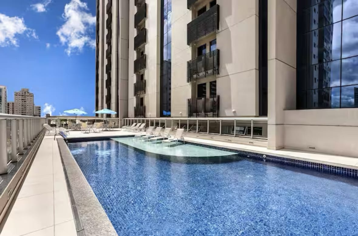 A sparkling outdoor pool is bordered by a spacious deck, featuring several lounge chairs and umbrellas. The building's reflective glass facade complements the clear blue sky, creating a serene environment for relaxation among the city landscape.