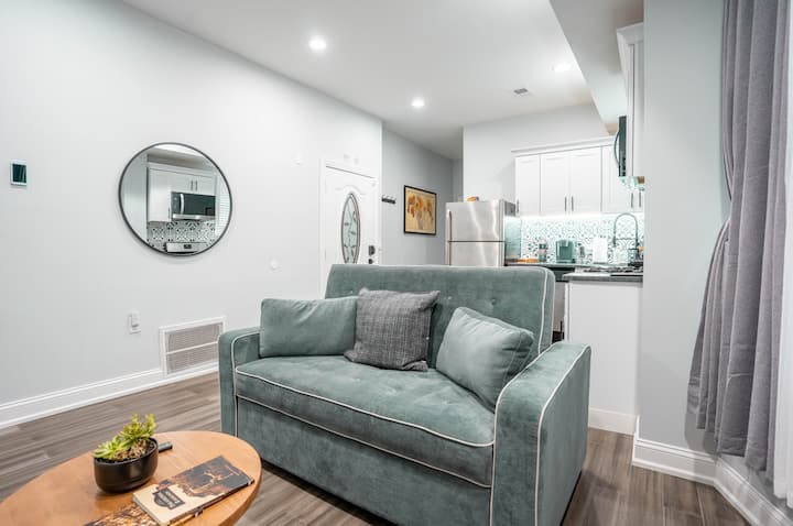 The Loft at Cedar Park living room features a stylish teal sofa sleeper, a modern round coffee table, and an open view into the bright, updated kitchen.