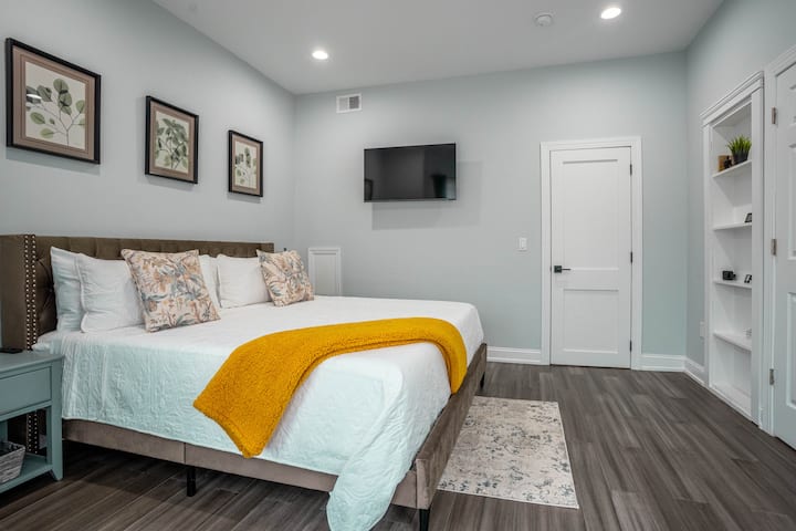 The Loft at Cedar Park King Bedroom features a luxurious bed, wall mounted TV, and built in shelving. Clean lines and neutral tones are accented by pops of yellow for a warm, modern feel.