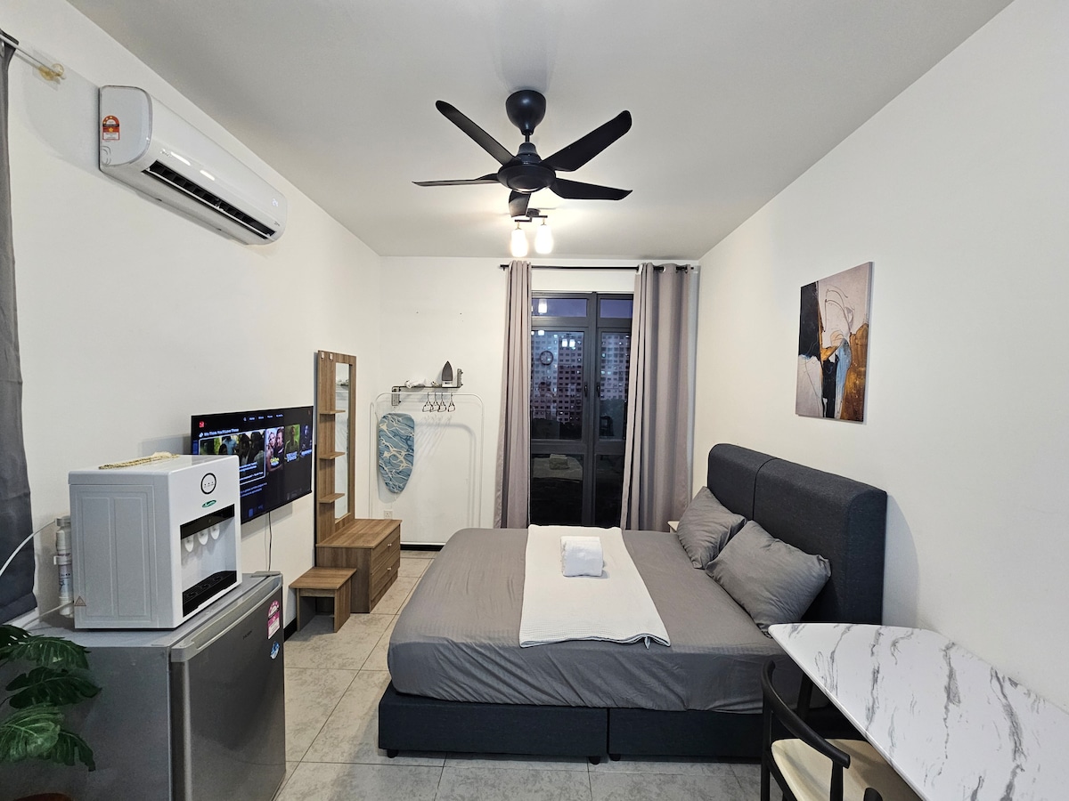 A private studio with a spacious bed covered in a white sheet, positioned near a window. A ceiling fan and wall-mounted television are present. A small kitchenette is available, featuring a refrigerator and a marble-top table. Light curtains frame the window, allowing natural light to enter.