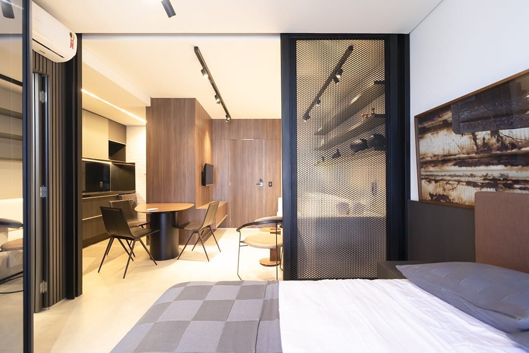 A stylish living space is visible, featuring a clear division between the sleeping area and the dining space. A spacious bed is positioned in the foreground, while a circular dining table and black chairs are seen through the glass partition. Warm wood accents add to the modern aesthetic.