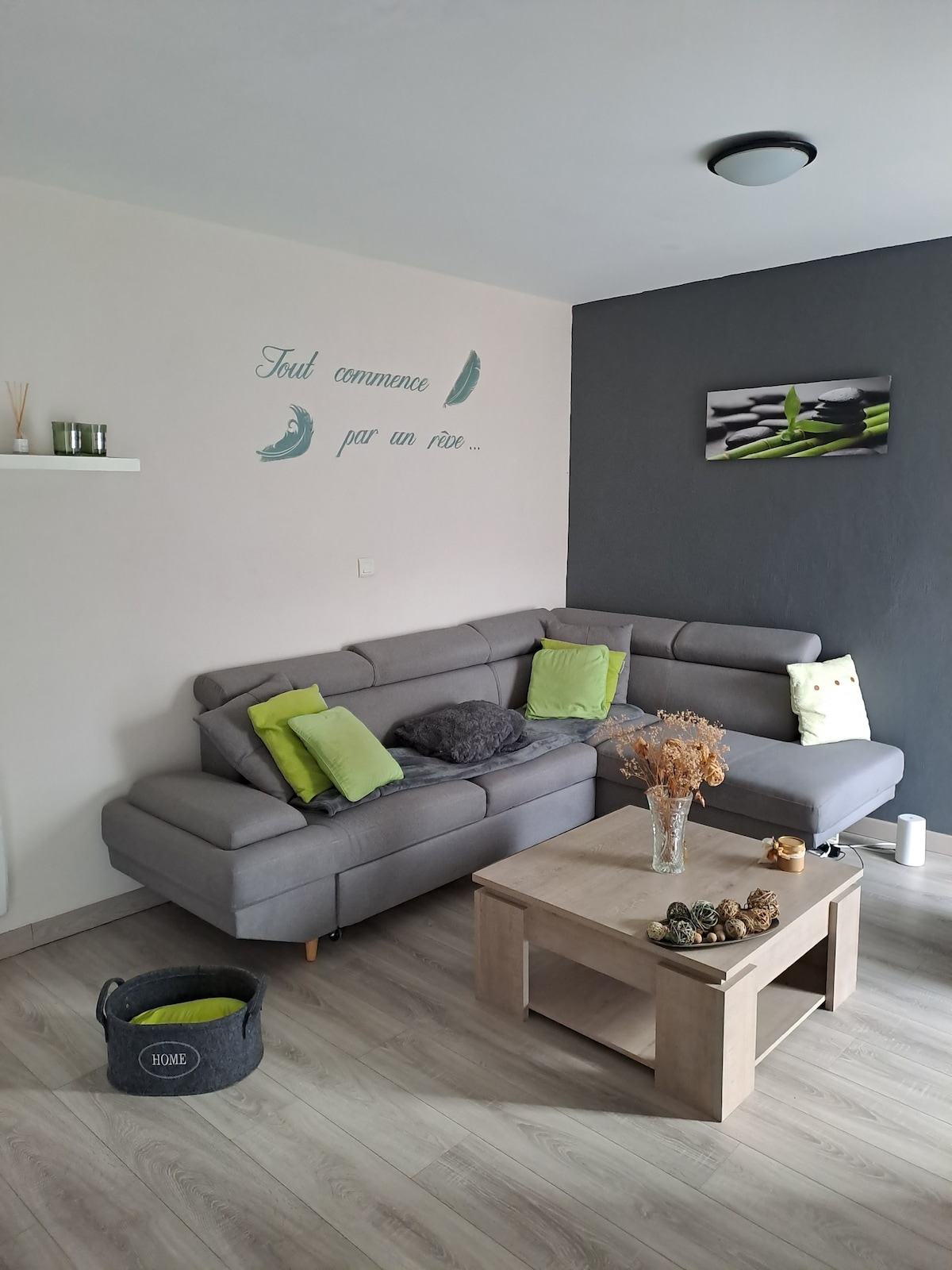 A spacious living area is furnished with a modern grey sectional sofa accented by green cushions. A wooden coffee table, adorned with decorative elements, sits at the center. A wall is decorated with a simple phrase, and a piece of artwork complements the atmosphere.