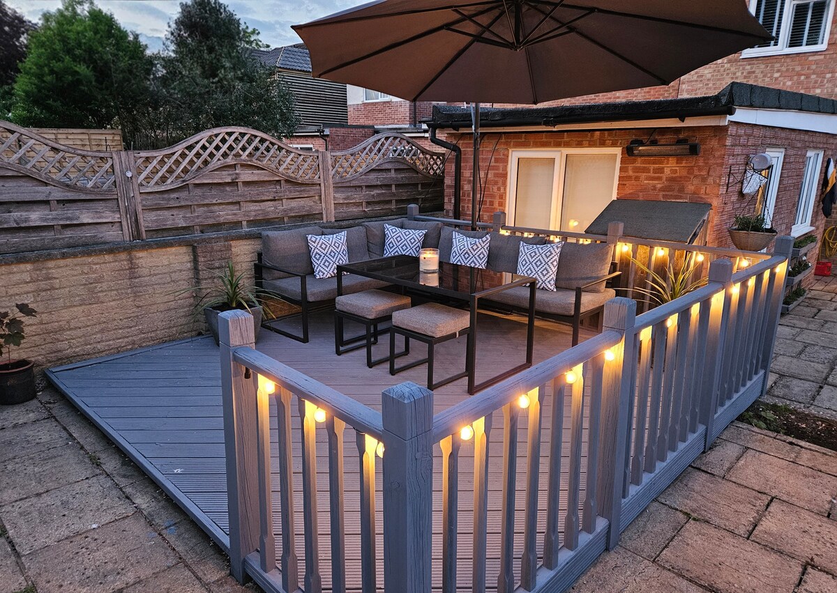 An outdoor seating area is presented with modern furniture, featuring a table surrounded by cushioned chairs. Soft lighting illuminates the space, enhancing its welcoming appeal. A large umbrella provides shade, while the area is enclosed by a grey railing, ensuring a comfortable environment for relaxation.
