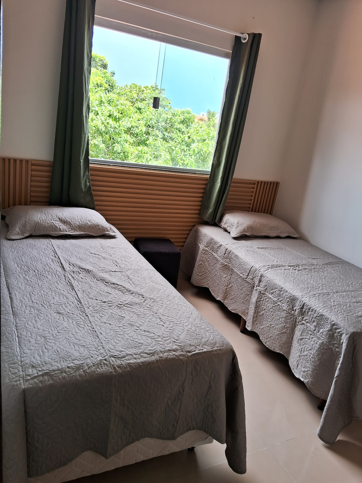 A bright sleeping area features two twin beds dressed in light grey bedding. Natural light floods through a large window, adorned with green curtains. A small bedside table sits between the beds, offering a minimalist and airy environment.