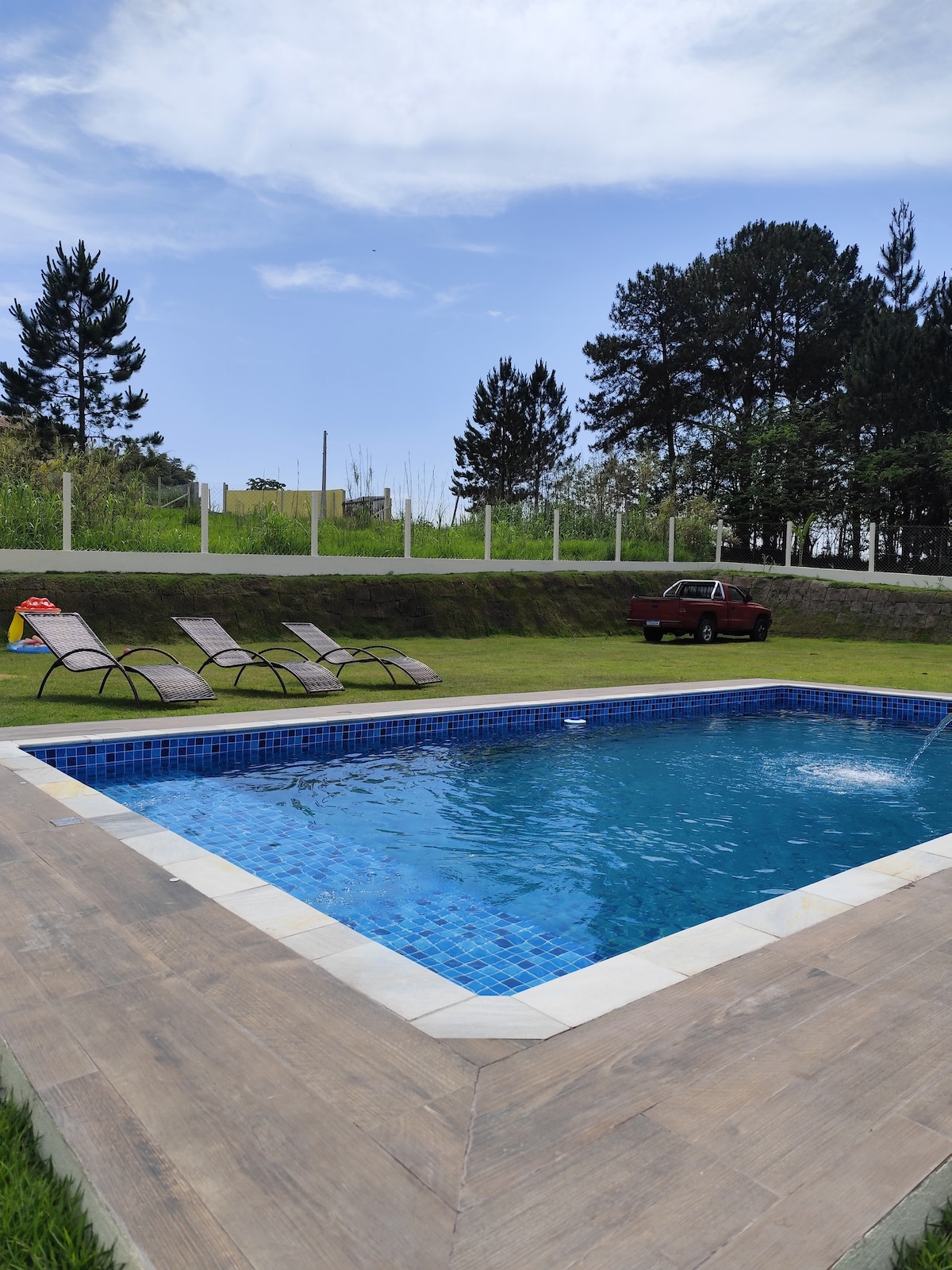 A clear swimming pool is surrounded by manicured green grass and lounge chairs, inviting relaxation. The pool features a mosaic tiled border and a gentle waterfall. In the background, tall trees frame the area, providing a sense of tranquility and nature.
