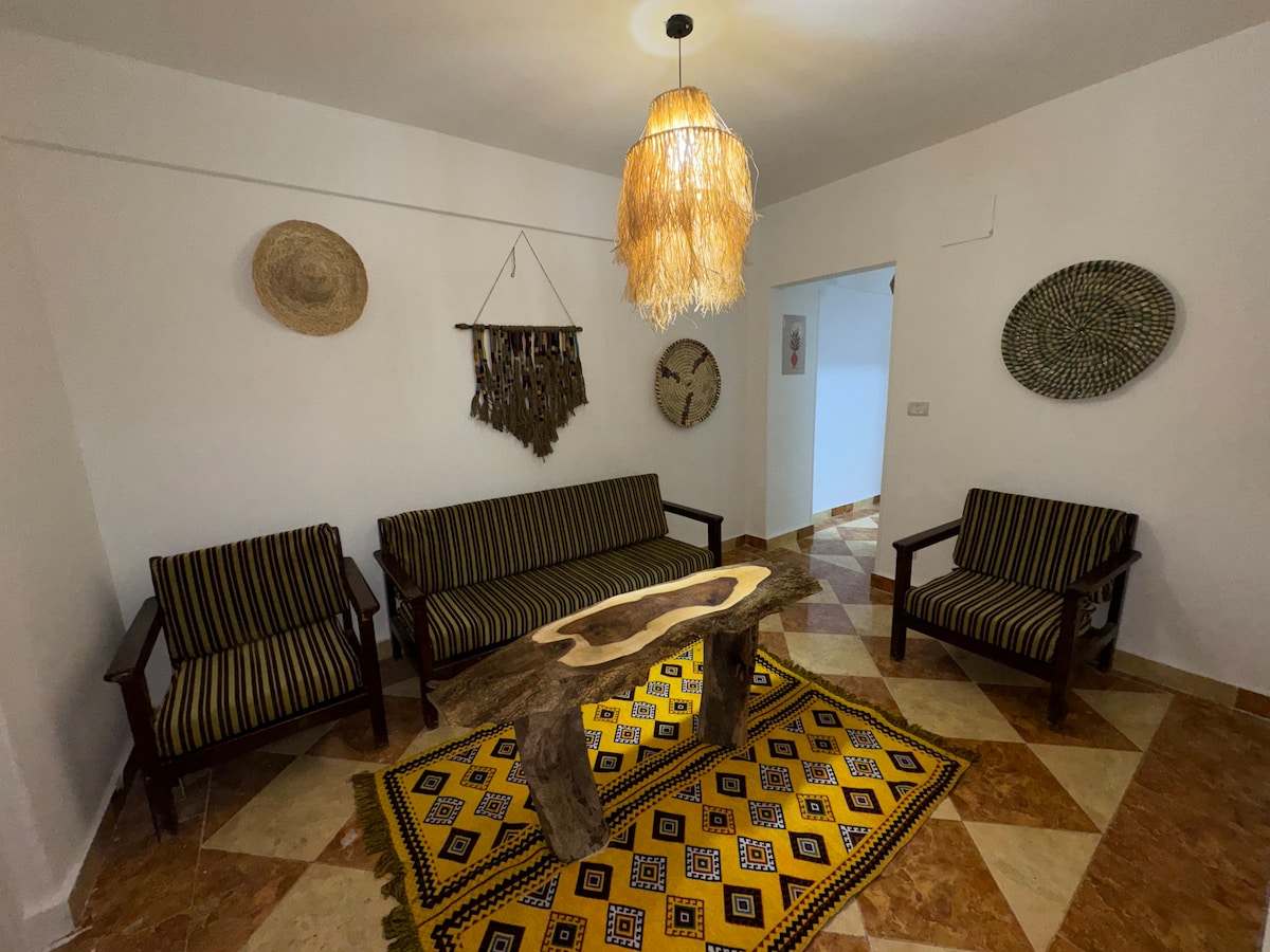 A welcoming sitting area features two striped armchairs and a matching sofa, arranged around a unique wooden coffee table. A traditional patterned rug defines the space, while woven wall hangings and a decorative clock contribute to the room's bohemian charm.