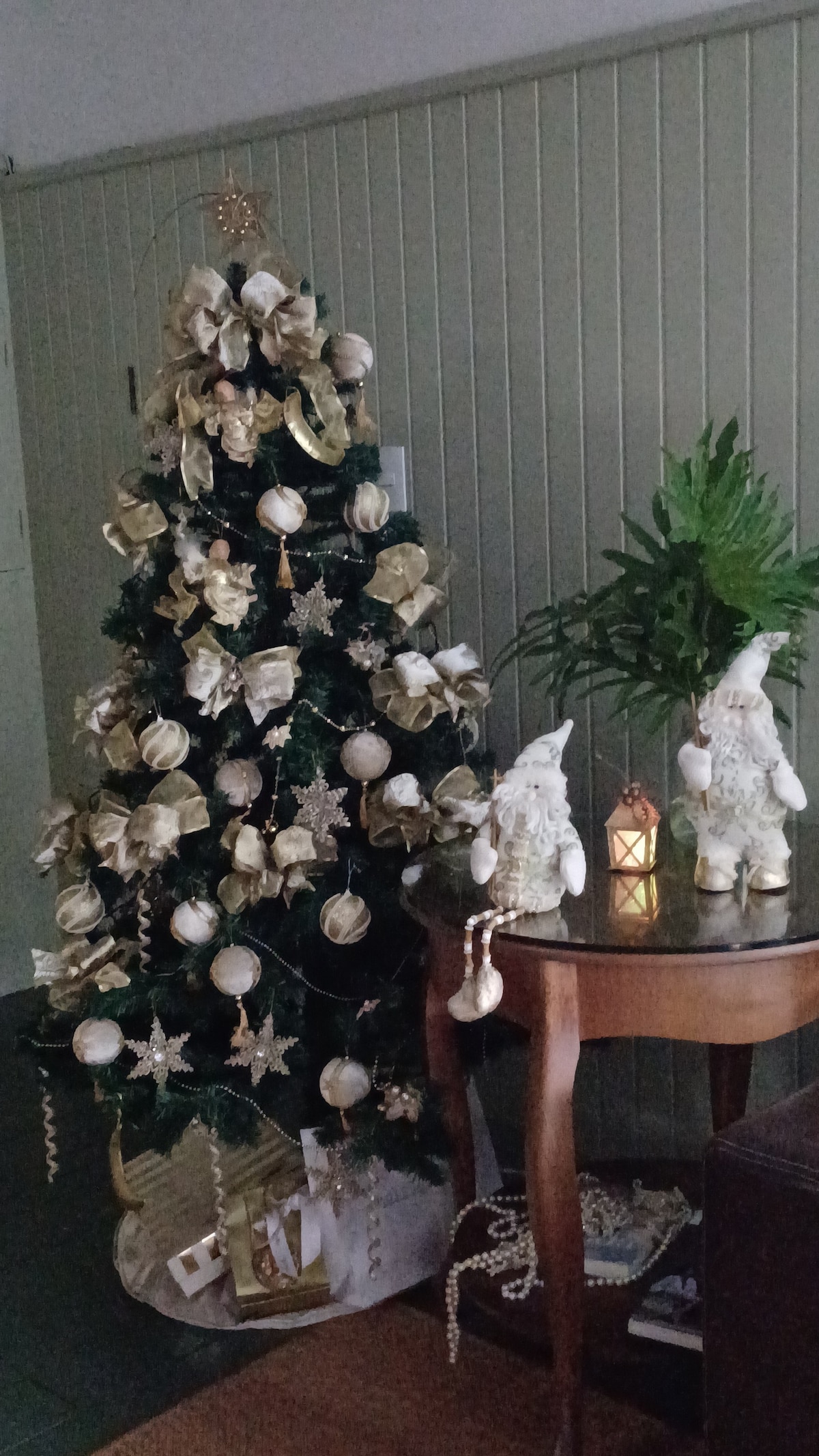 A decorated Christmas tree, adorned with golden ornaments and stars, stands near a small table. The table features a decorative lantern and two elegant figurines. Lush green leaves are displayed beside the tree, complementing the festive decor.