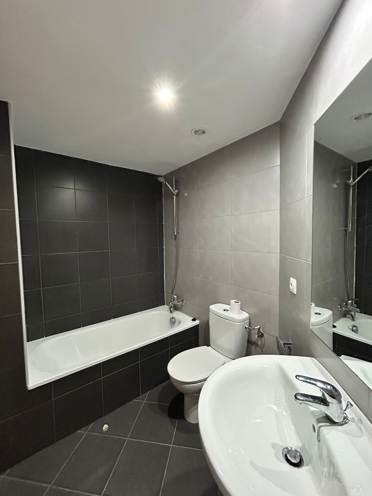The bathroom features a modern design with sleek black and gray tiles. A bathtub with a showerhead is positioned next to a contemporary toilet. A circular mirror is mounted above a white sink, reflecting soft lighting from the ceiling.