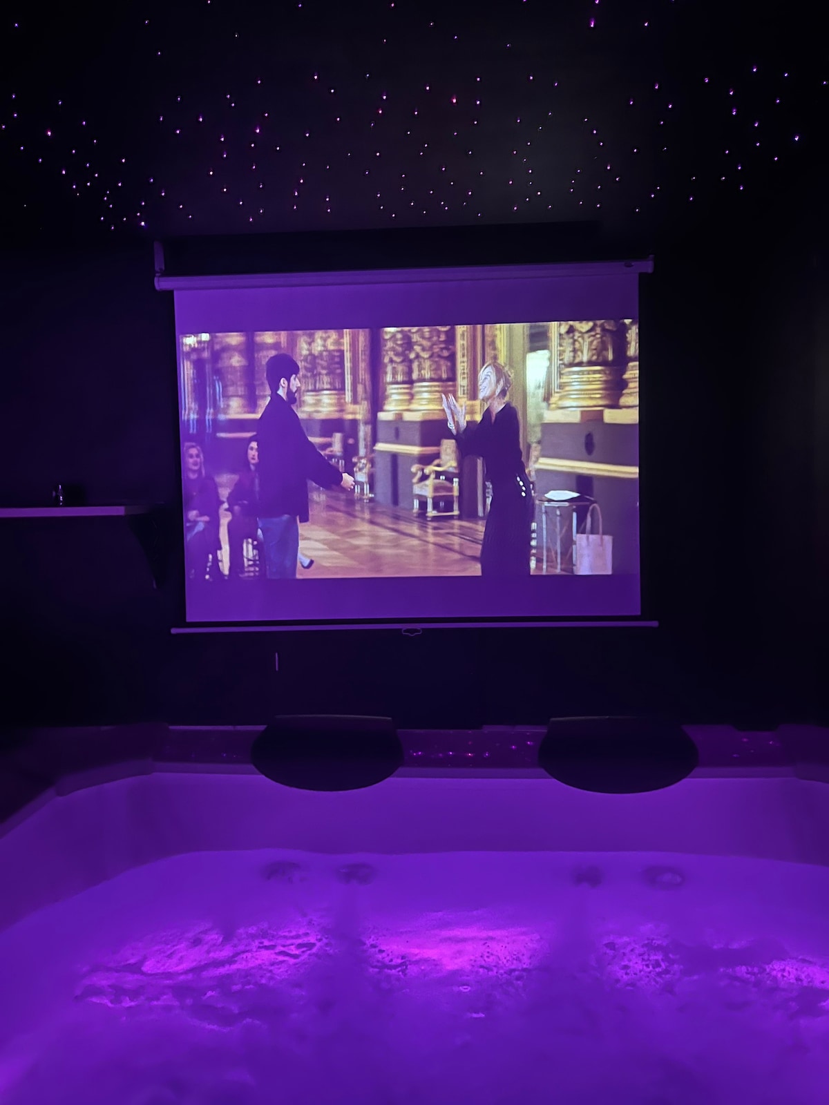 A jacuzzi spa is visible, illuminated with soft purple lighting. Above, a large screen displays a movie, enhancing the atmosphere. A starry night sky effect adorns the ceiling, creating a calming and immersive environment for relaxation.