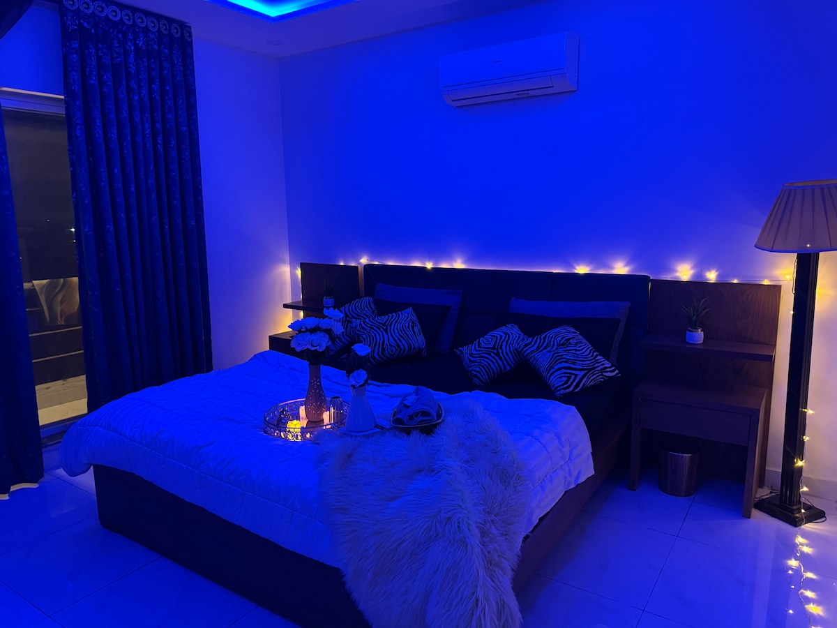 A serene bedroom is featuring a spacious bed adorned with soft linens and decorative pillows. Soft blue lighting and twinkling fairy lights enhance the tranquil ambiance. A large window offers access to a captivating view, while a stylish lamp and small plant add a touch of elegance.