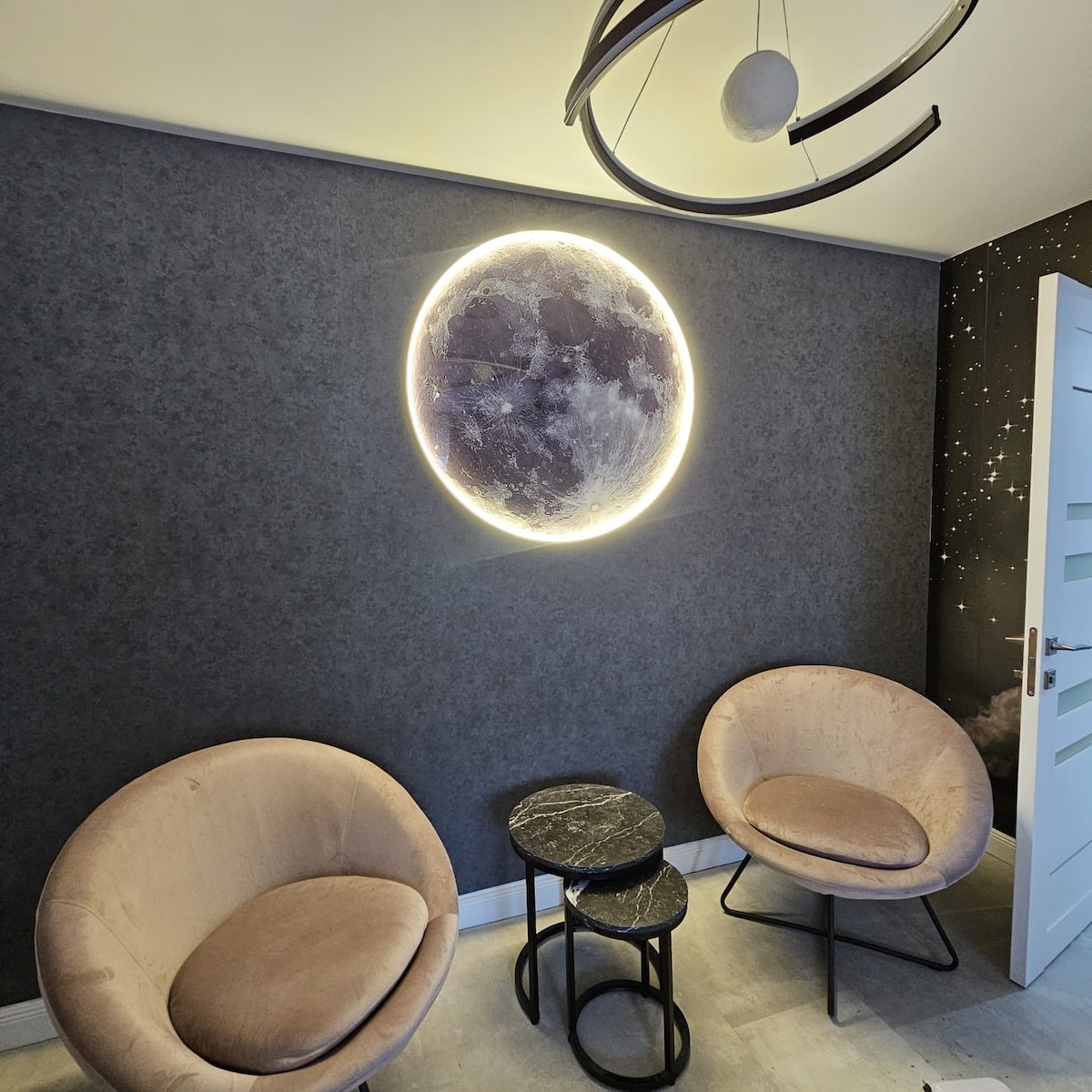 A cozy seating area features two round armchairs with a soft finish, positioned near a modern tabletop. A circular, illuminated moon art piece on the dark wall adds a calming focal point, while a sleek ceiling fixture completes the contemporary design.
