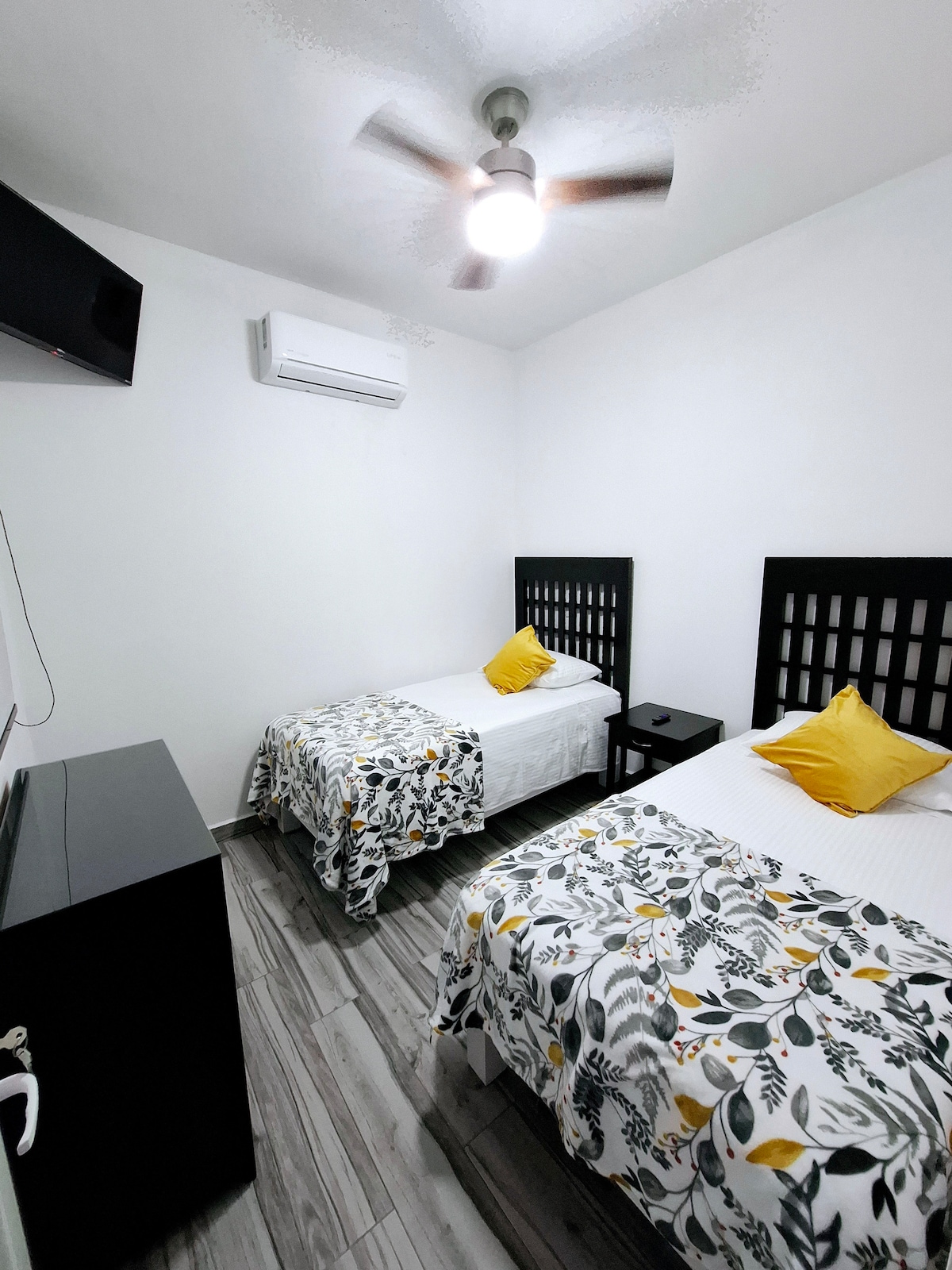 This room features two twin beds, each adorned with a floral-patterned blanket and a bright yellow accent pillow. A small nightstand is positioned between the beds, and a wall-mounted television is visible. An air conditioning unit and ceiling fan contribute to the comfort of the space.