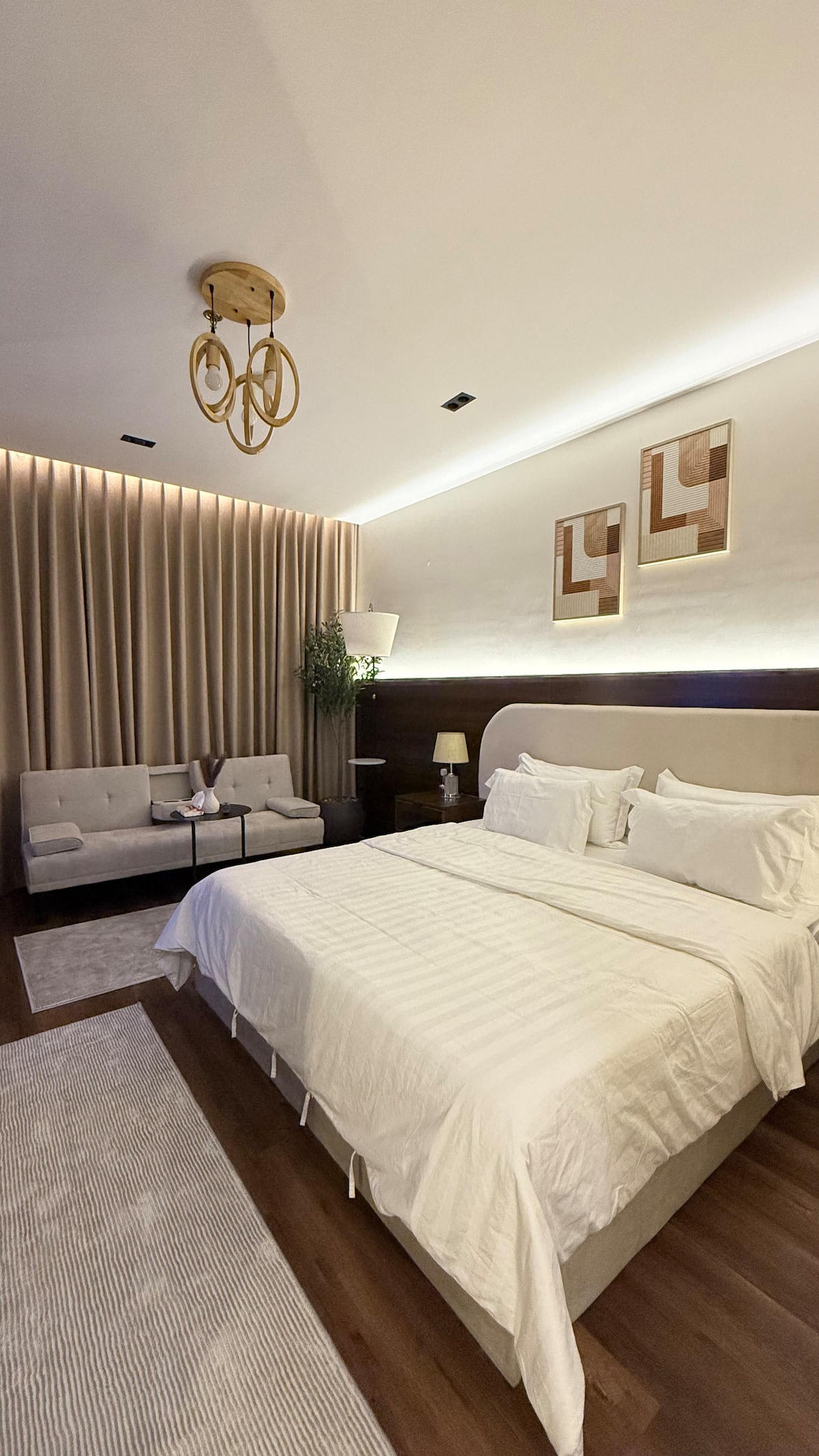 A spacious bedroom features a large bed dressed in crisp white linens. Soft lighting highlights the textured wall behind the bed, while two abstract art pieces hang above. A cozy seating area with a small table is positioned beside the bed, surrounded by warm tones and a neutral rug.