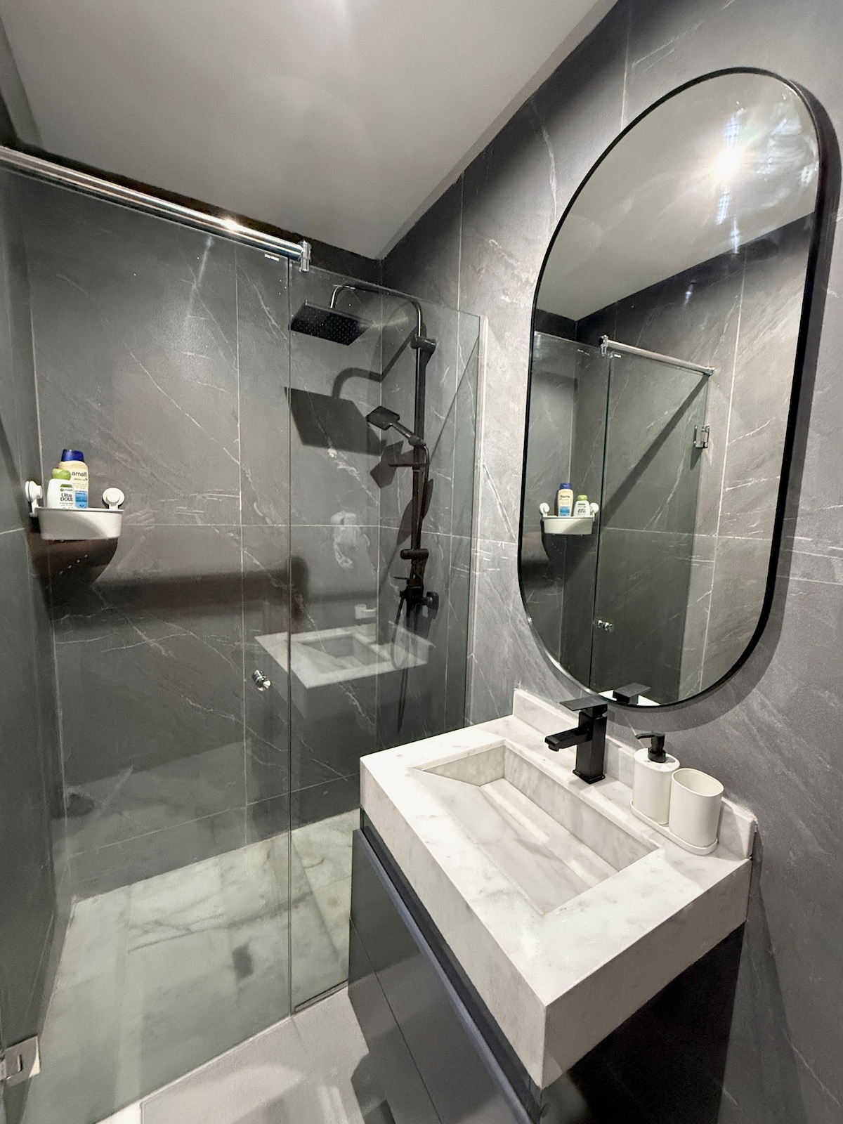A modern bathroom is presented, featuring sleek gray marble walls and flooring. A spacious walk-in shower is equipped with a rain showerhead. A minimalist vanity with a white marble sink is accompanied by a round mirror, complemented by toiletries neatly arranged on shelves.