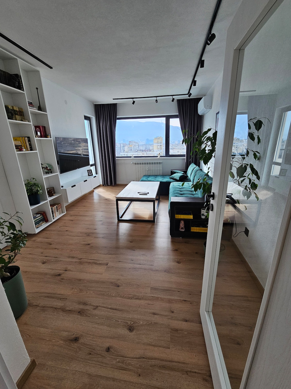 A bright and airy living space is presented, featuring a comfortable green sofa and a minimalist coffee table. Large windows provide panoramic views of the city skyline, and natural light enhances the warm wooden flooring. Shelving units display books and decorative plants along the walls.