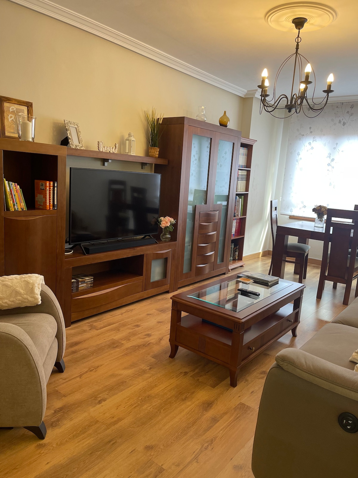 A cozy living area features a wooden entertainment unit with a flat-screen TV and a glass coffee table. A dining table is positioned nearby, surrounded by chairs. Natural light enters through a window adorned with light curtains, enhancing the warm ambiance of the space.