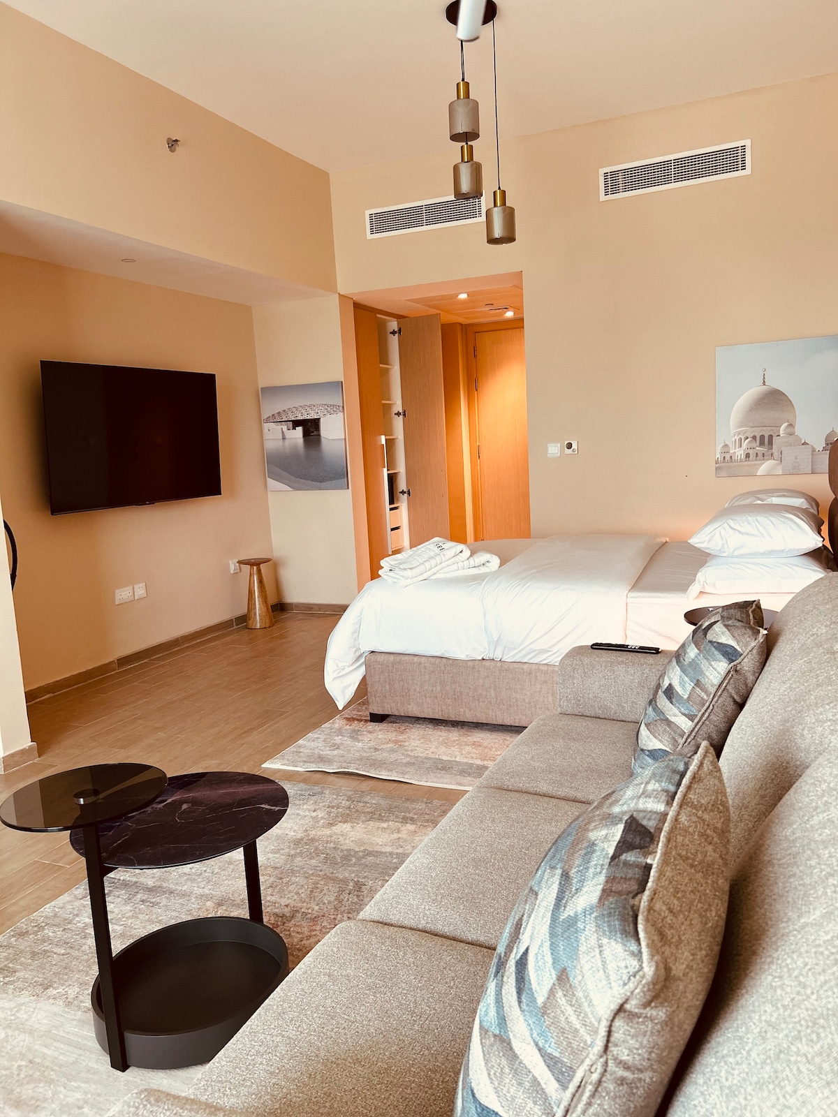 A modern studio features a spacious bed and a comfortable sofa positioned near a sleek coffee table. A large flat-screen television is mounted on the wall, and light wood accents create a warm ambiance. Soft lighting enhances the space, while an artistic piece adds character.