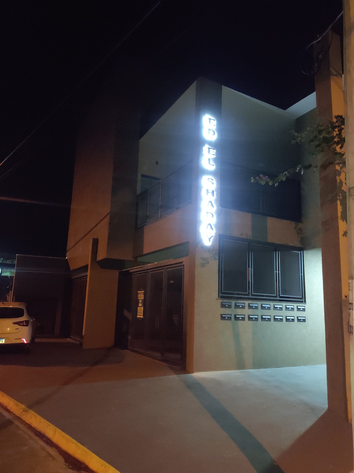 The exterior of a modern building is illuminated at night, featuring a prominent sign displaying the property name. Nearby mailboxes are visible alongside the entrance, bordered by a well-maintained walkway. The surrounding area is lined with soft street lighting, enhancing visibility.