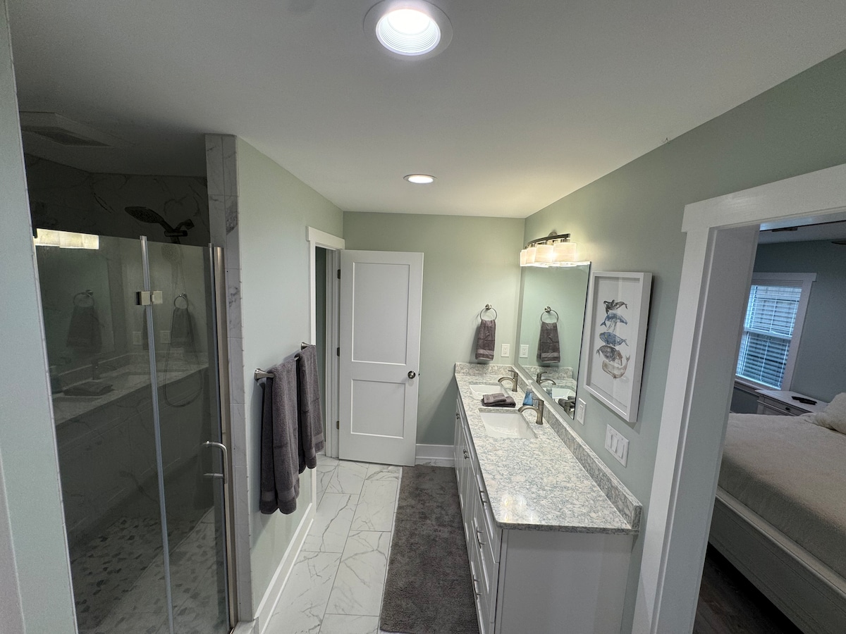 A modern bathroom featuring a spacious double vanity with granite countertops and decorative wall art. A glass shower enclosure is visible to the left, while plush towels hang neatly on the wall. Soft lighting complements the pale green walls, creating a fresh and clean atmosphere.