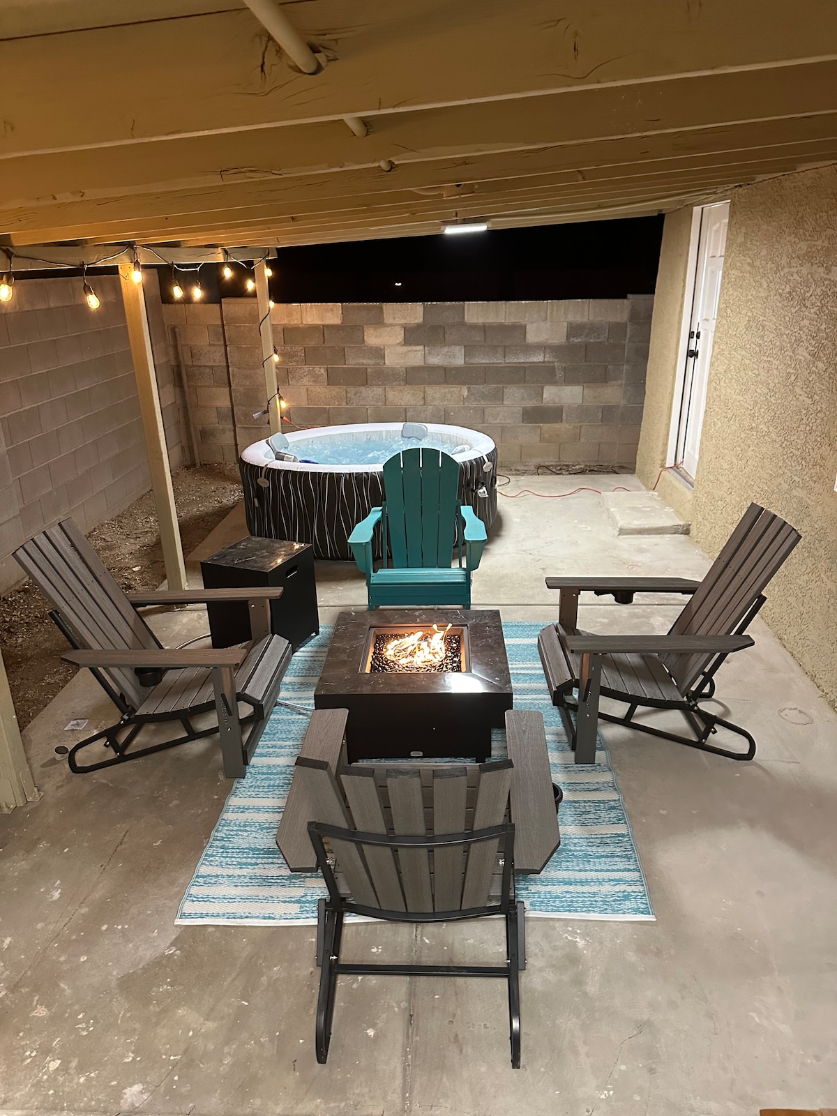A cozy outdoor seating area features four Adirondack chairs arranged around a central fire pit. A soft blue and white rug lies beneath the seating, creating a comfortable atmosphere. A hot tub is visible in the background, complemented by string lights overhead.