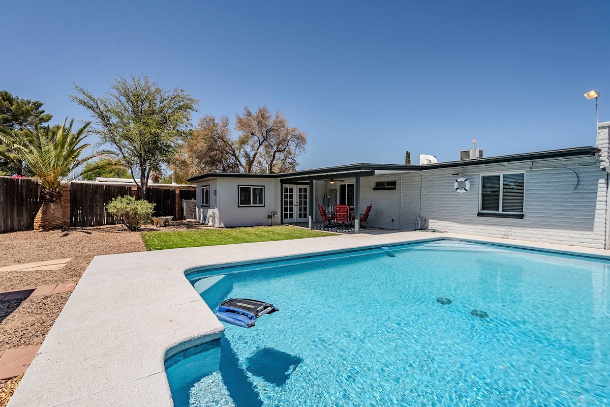 A clear view of the renovated home and pool area shows a spacious backyard featuring a large, inviting swimming pool. Surrounding greenery, including palm trees, lines the yard, while comfortable seating options are positioned near the entrance of the house.