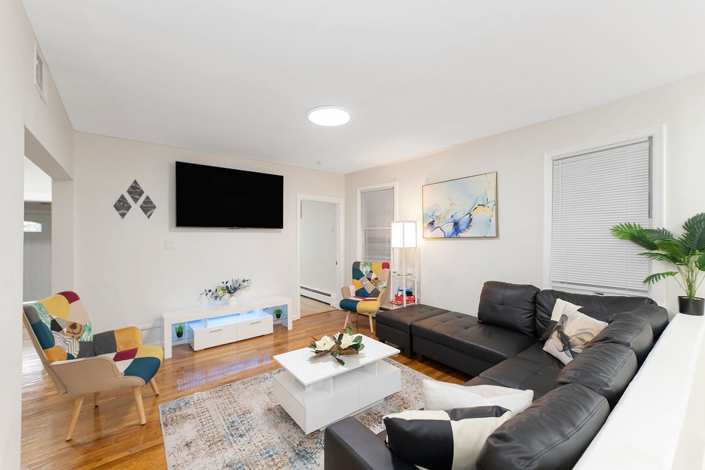The living room features a sectional couch and two accent chairs, arranged around a modern coffee table. A large wall-mounted TV is positioned above a stylish console. Natural light filters through the windows, and a vibrant area rug adds warmth to the hardwood floor.