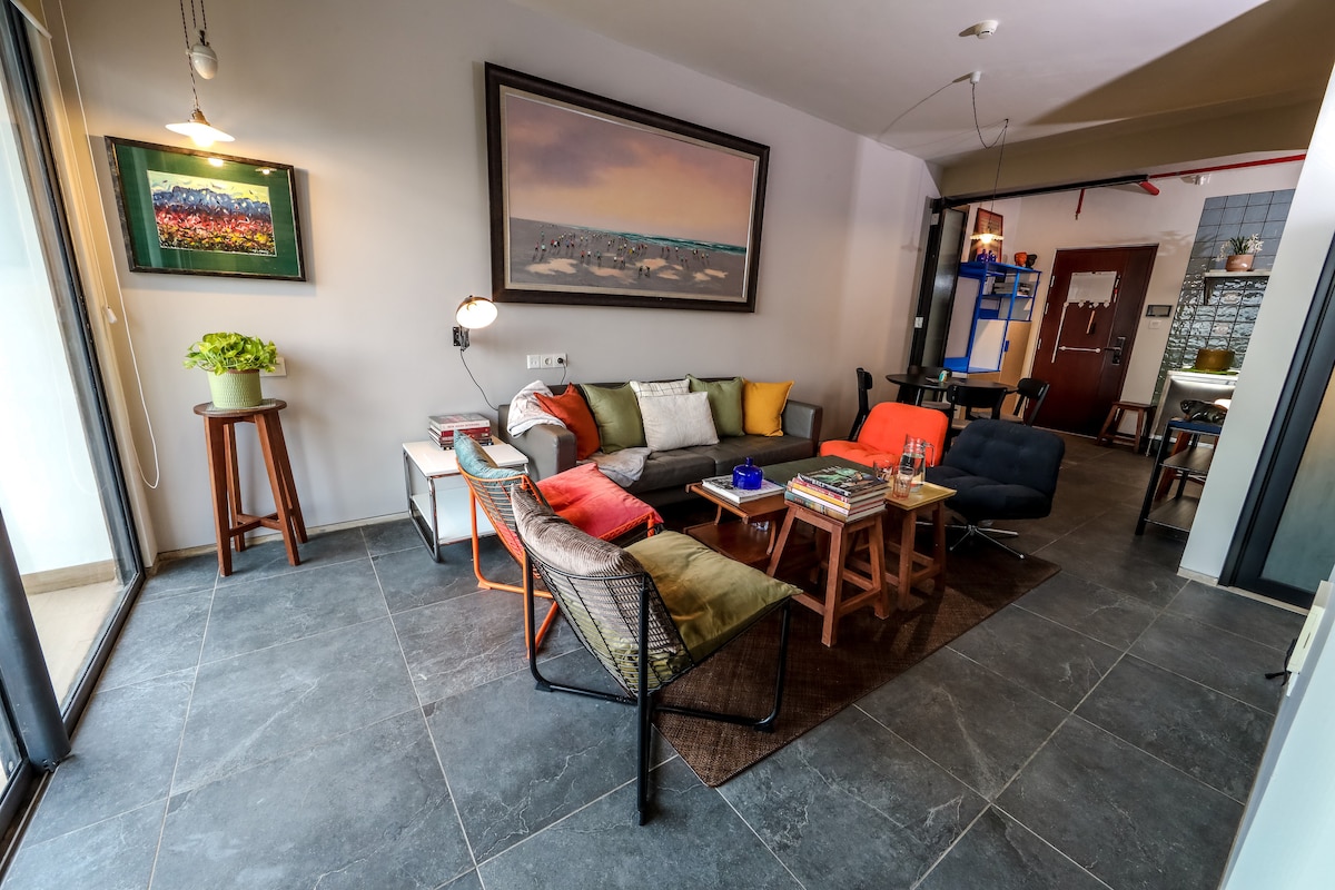 A comfortable living room features a mix of seating options, including a large, colorful sofa and mid-century armchairs. Natural light streams through a sliding glass door, illuminating the stone-tiled floor. Decorative elements include a framed artwork and a small potted plant on a side table.