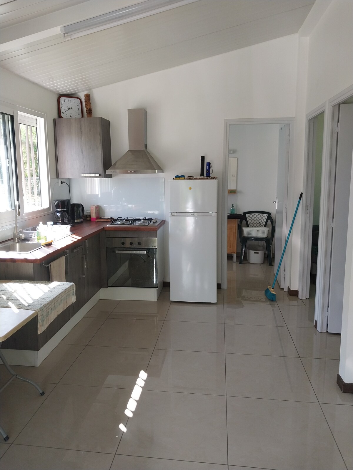 The kitchen features modern appliances, including a stainless steel refrigerator and a gas stove. A small dining table is situated near large windows, allowing natural light to fill the space. Neutral-colored cabinets complement the tiled floor, adding to the room's clean and functional design.