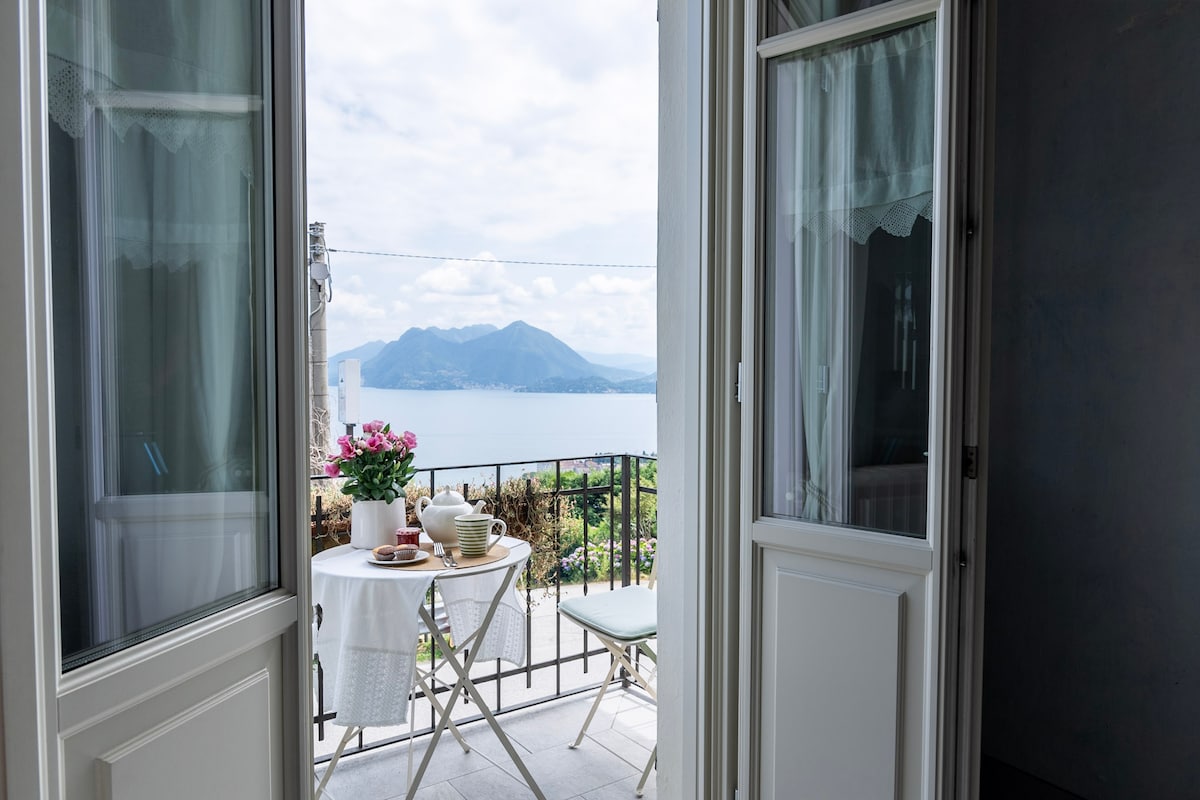 An outdoor balcony is accessed through open double doors, featuring a small table set for breakfast with a vase of flowers. The scenic view includes mountains and a calming body of water, inviting relaxation amidst a serene landscape.