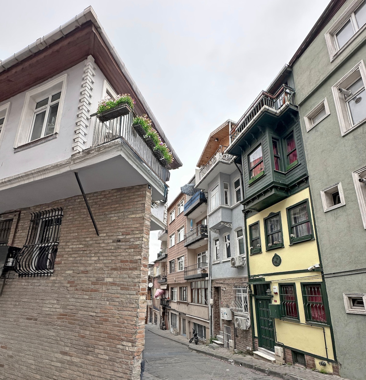A narrow street is framed by colorful buildings featuring traditional architecture. A balcony adorned with potted plants is visible on the left, while a variety of window designs and façades showcase the neighborhood's charm on the right.