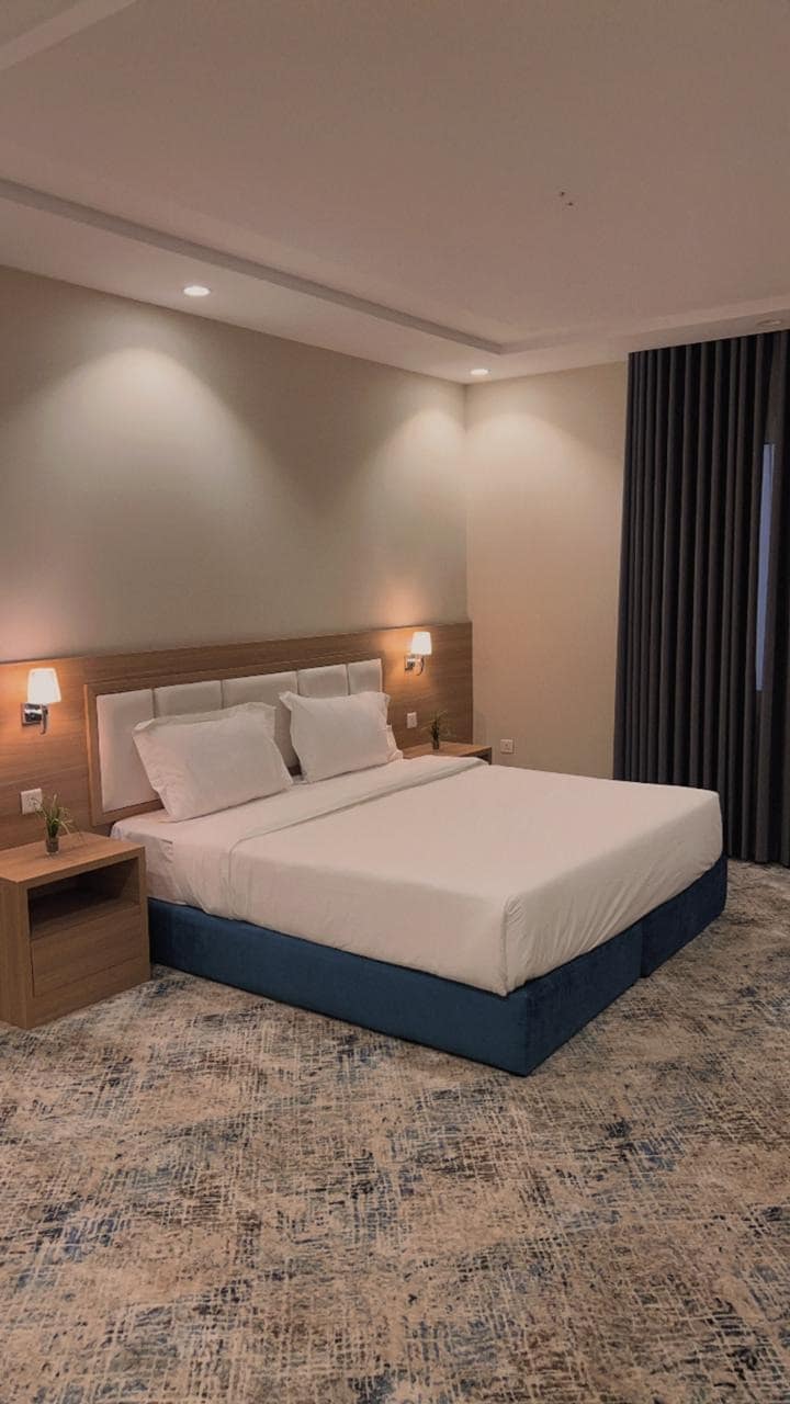 A well-lit bedroom features a spacious bed with crisp white linens and two nightstands on either side. Soft gray curtains frame the window, while a patterned area rug adds texture to the floor. Subtle wall sconces provide ambient lighting.