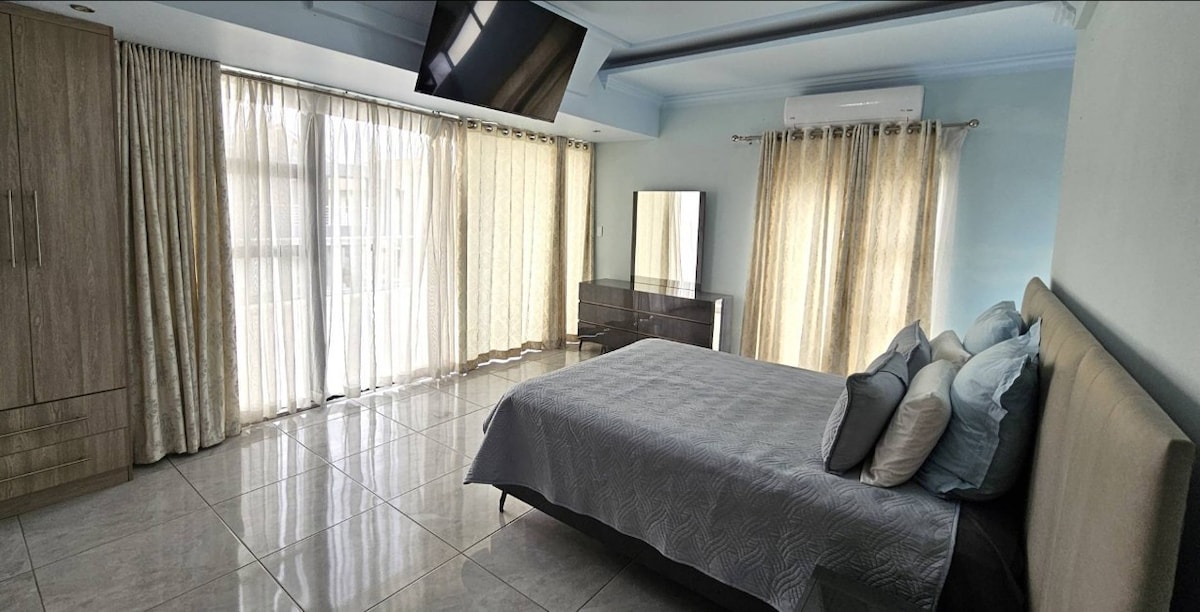 A spacious bedroom features large windows covered with sheer curtains, allowing natural light to fill the room. A comfortable bed is dressed in light grey linens, accompanied by several decorative pillows. A sleek mirror and modern furniture create a minimalist aesthetic.