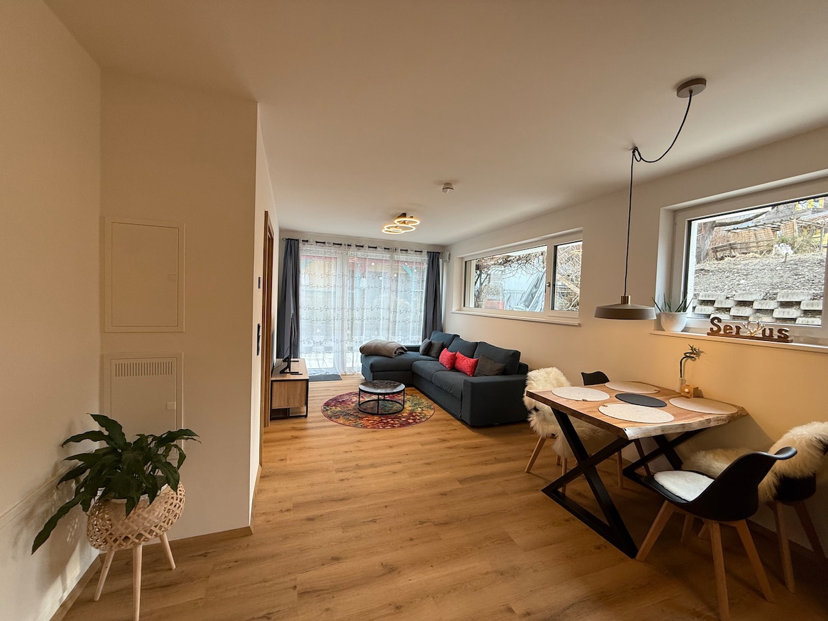 The living and dining area features modern furnishings, including a dark blue sofa with decorative pillows and a round dining table with stylish chairs. Large windows allow natural light to brighten the space, and a colorful rug adds a warm touch to the wooden floor.