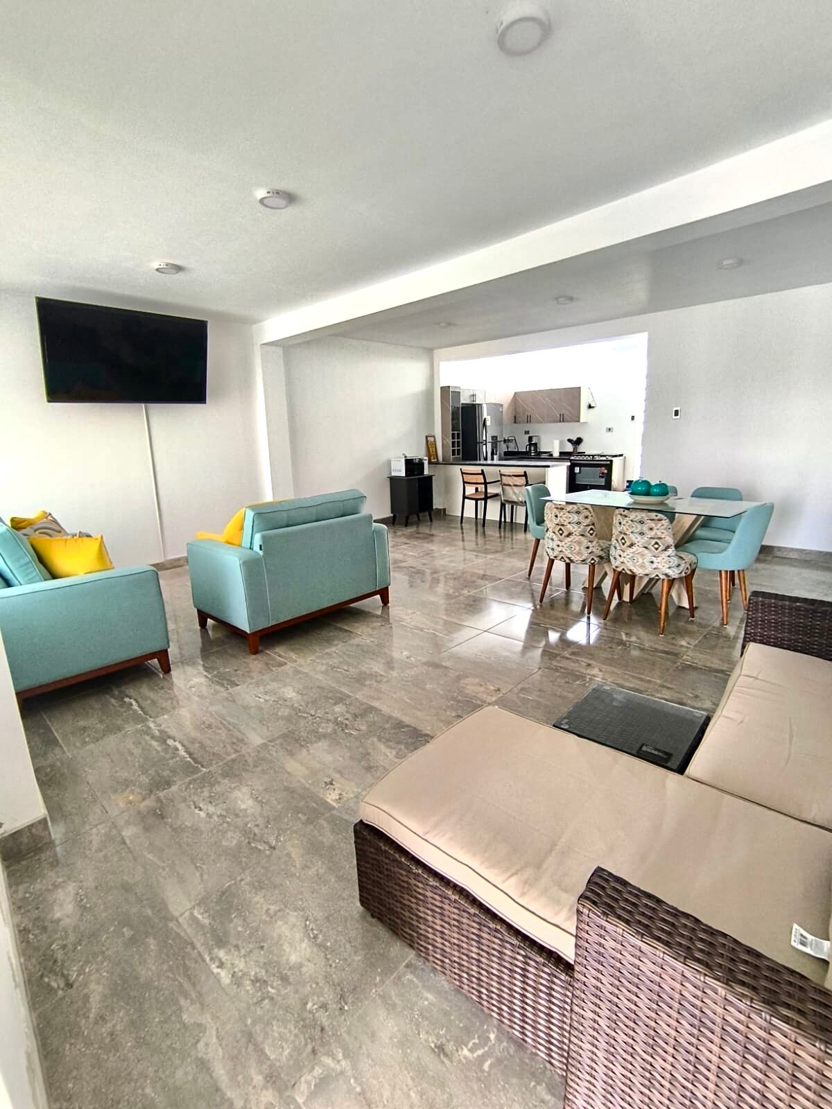 An open living area showcases a combination of light blue and woven seating options, complemented by a small dining table with chairs. A sleek television is mounted on the wall, and the space features modern, polished tile flooring. A kitchen area is visible in the background.