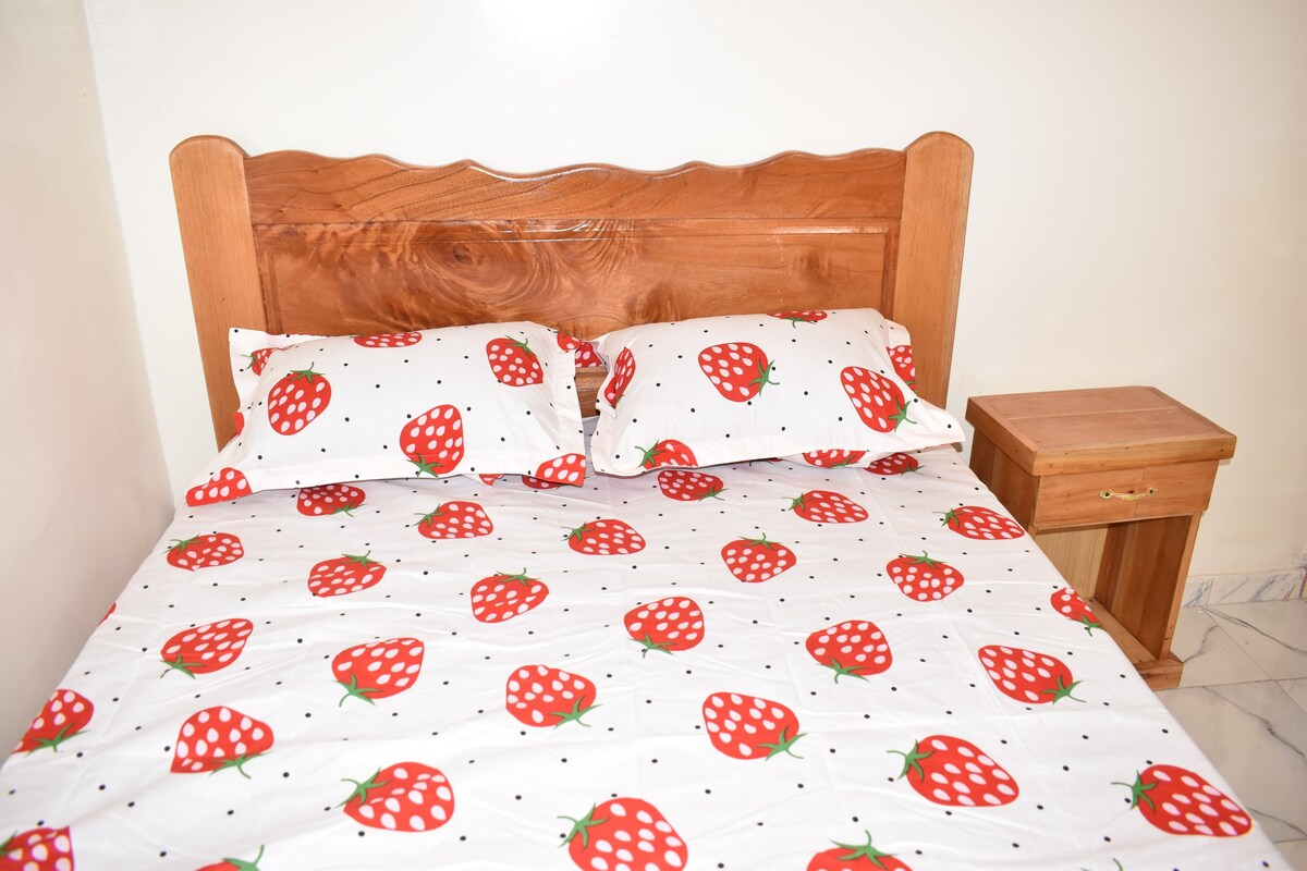 A spacious bed is dressed in a playful strawberry-patterned duvet cover and matching pillowcases. A wooden bedside table stands beside the bed, offering a simple and functional touch to the cozy sleeping area.