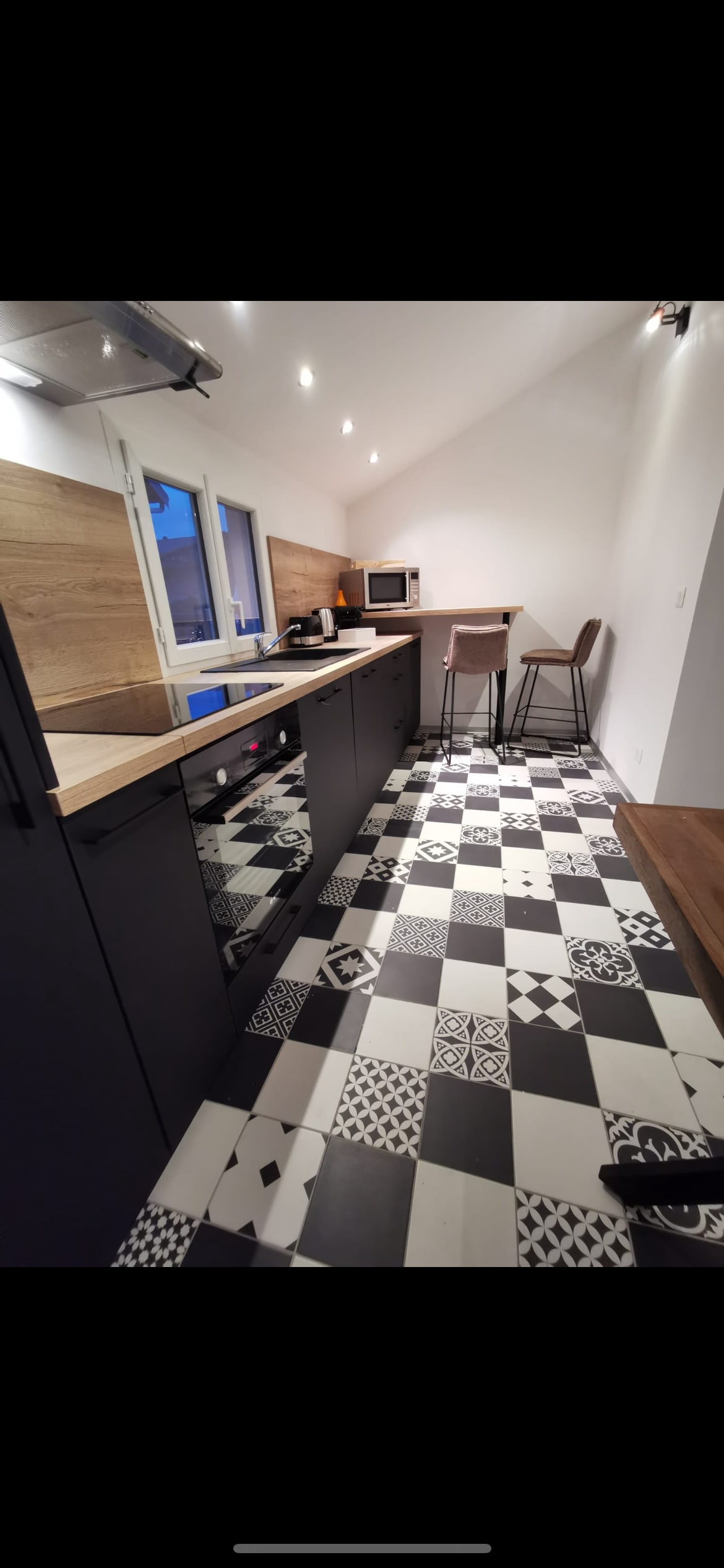 A modern kitchen is shown featuring a sleek black cabinetry design and a wooden countertop. The floor displays a distinct pattern of black and white tiles. A small dining area with two high bar stools is located near a window, providing natural light to the space.