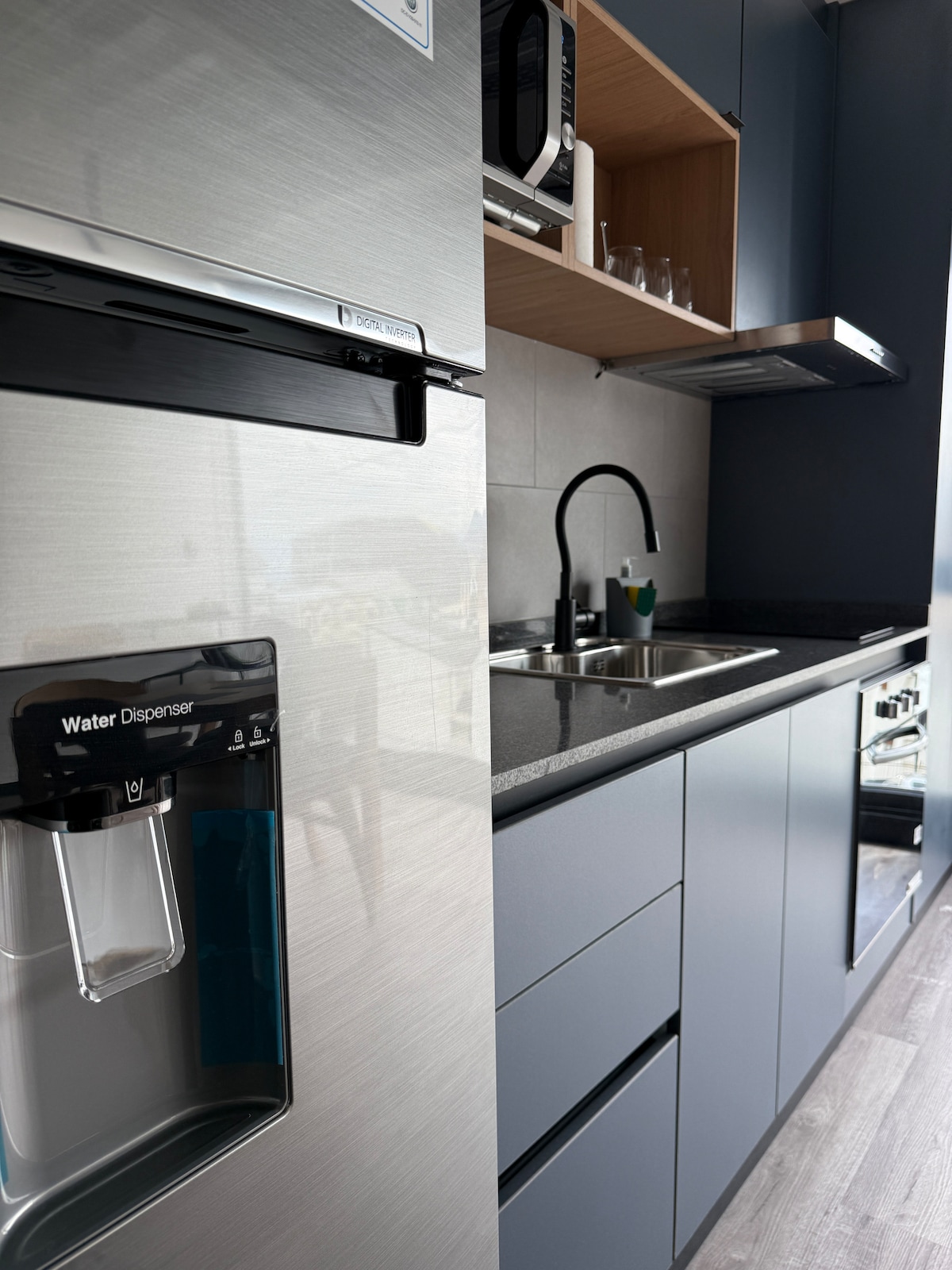 A modern kitchen area is shown, featuring a sleek stainless steel refrigerator with a water dispenser. A black sink is positioned next to a minimalist countertop, complemented by cabinetry in a neutral color palette. Above, kitchen appliances are neatly arranged, enhancing the functional space.