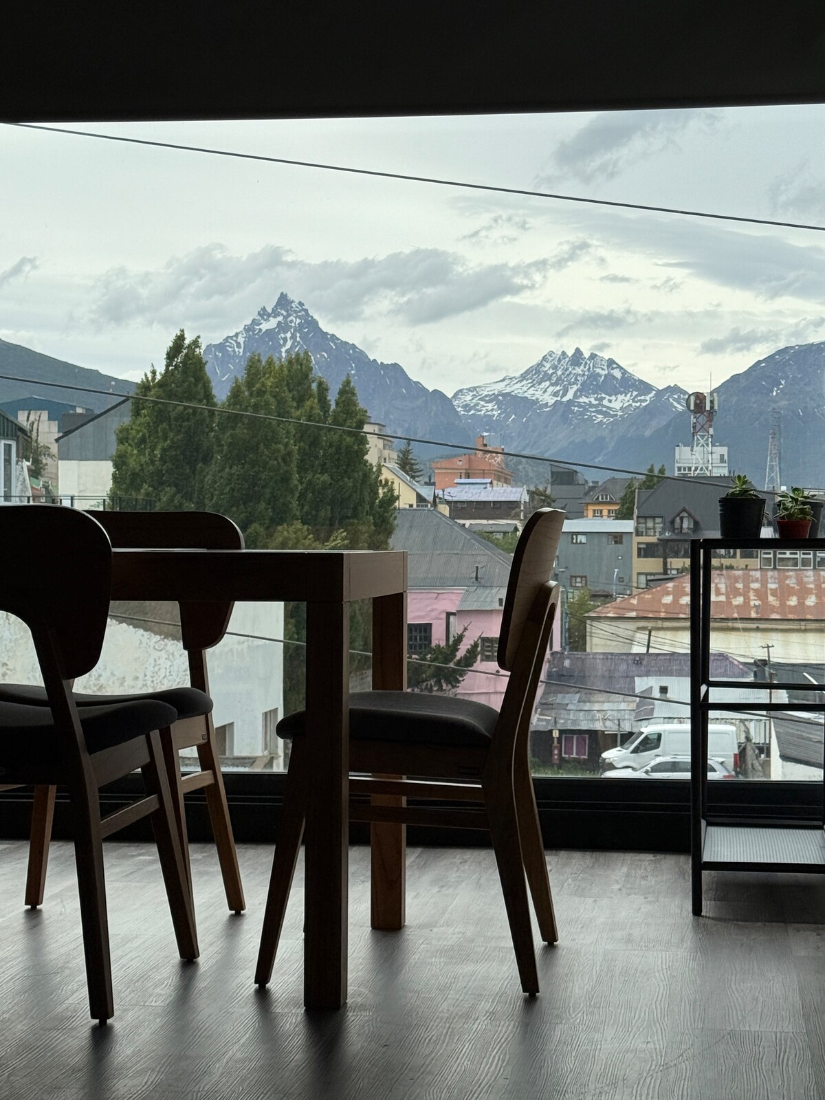 A dining table with wooden legs and black chairs is positioned beside a large window, offering a view of snow-capped mountains in the distance. The scene includes surrounding buildings and greenery, creating a contrasting urban and natural landscape.