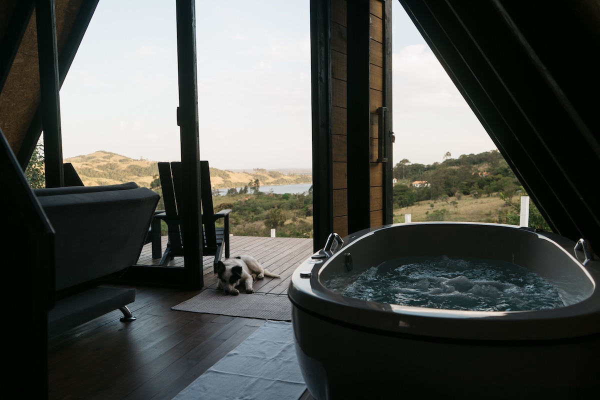 A double hydro-massage bathtub is positioned beside a large glass door, offering views of the lake and mountains. An inviting outdoor deck features a pair of chairs, while a cozy sofa sits nearby. Natural light fills the space, enhancing the serene atmosphere.