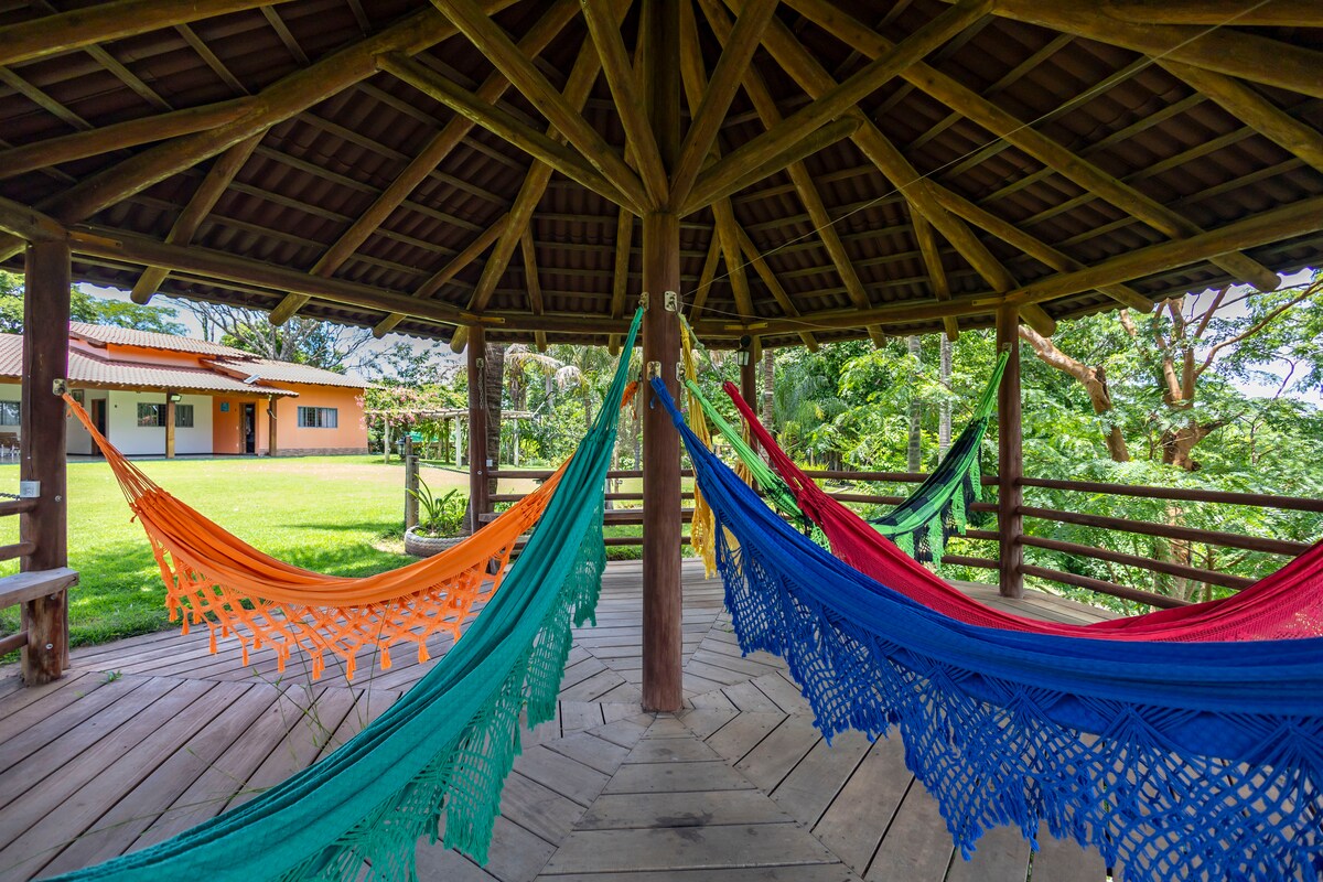 A spacious pavilion features eight colorful hammocks suspended in a circular arrangement, inviting relaxation. Wooden beams support a high ceiling, and a wooden deck surrounds the hammocks. Lush greenery is visible in the background, along with the facade of the house peeking through.