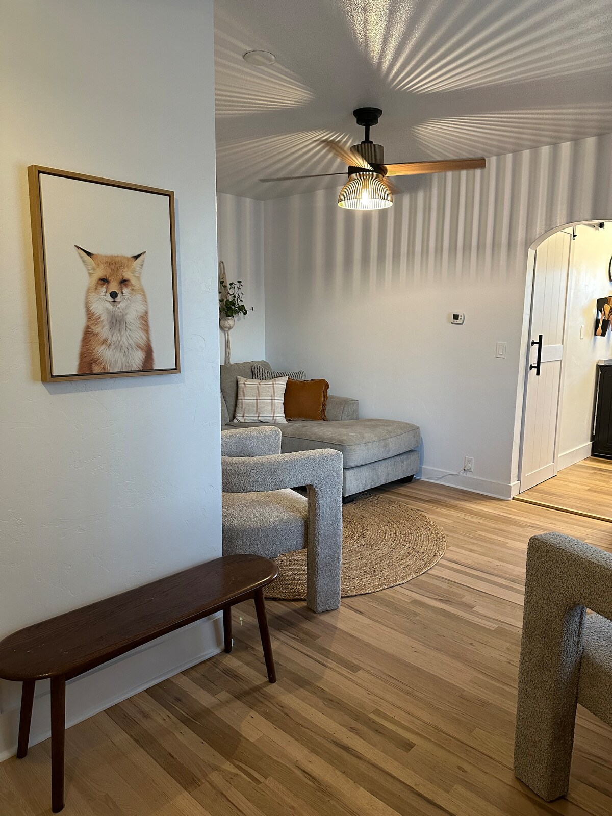 A comfortable living area features a light-colored sectional sofa with decorative cushions. A small round rug is placed under a wooden coffee table, and a fox portrait adorns the wall. Soft lighting creates gentle shadows across the space.