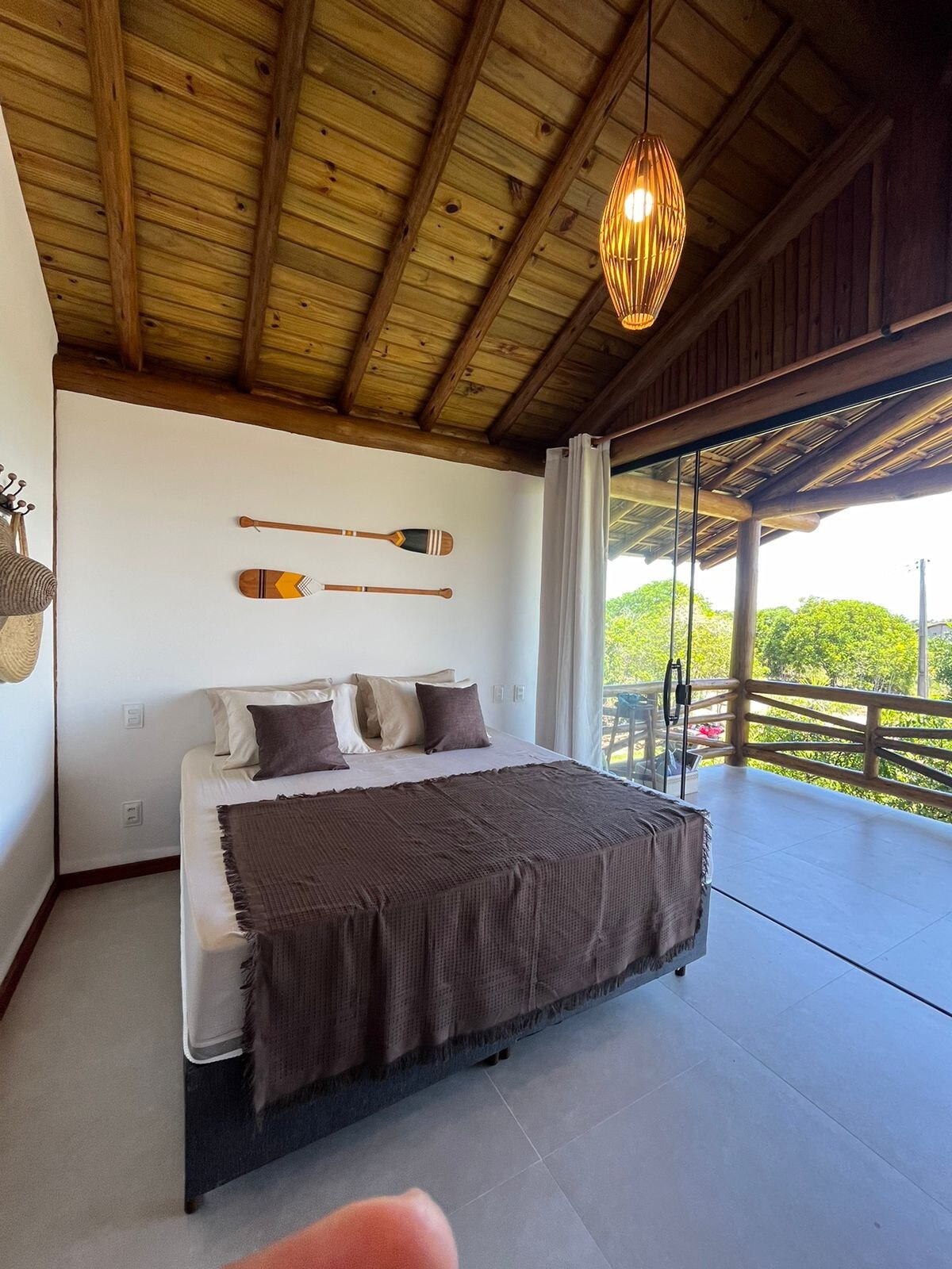 A spacious bedroom features a large bed adorned with decorative pillows, set against a backdrop of natural wood accents. Complementary wall art of paddles enhances the decor, while large glass doors allow natural light and provide access to a private balcony with views of the surrounding greenery.
