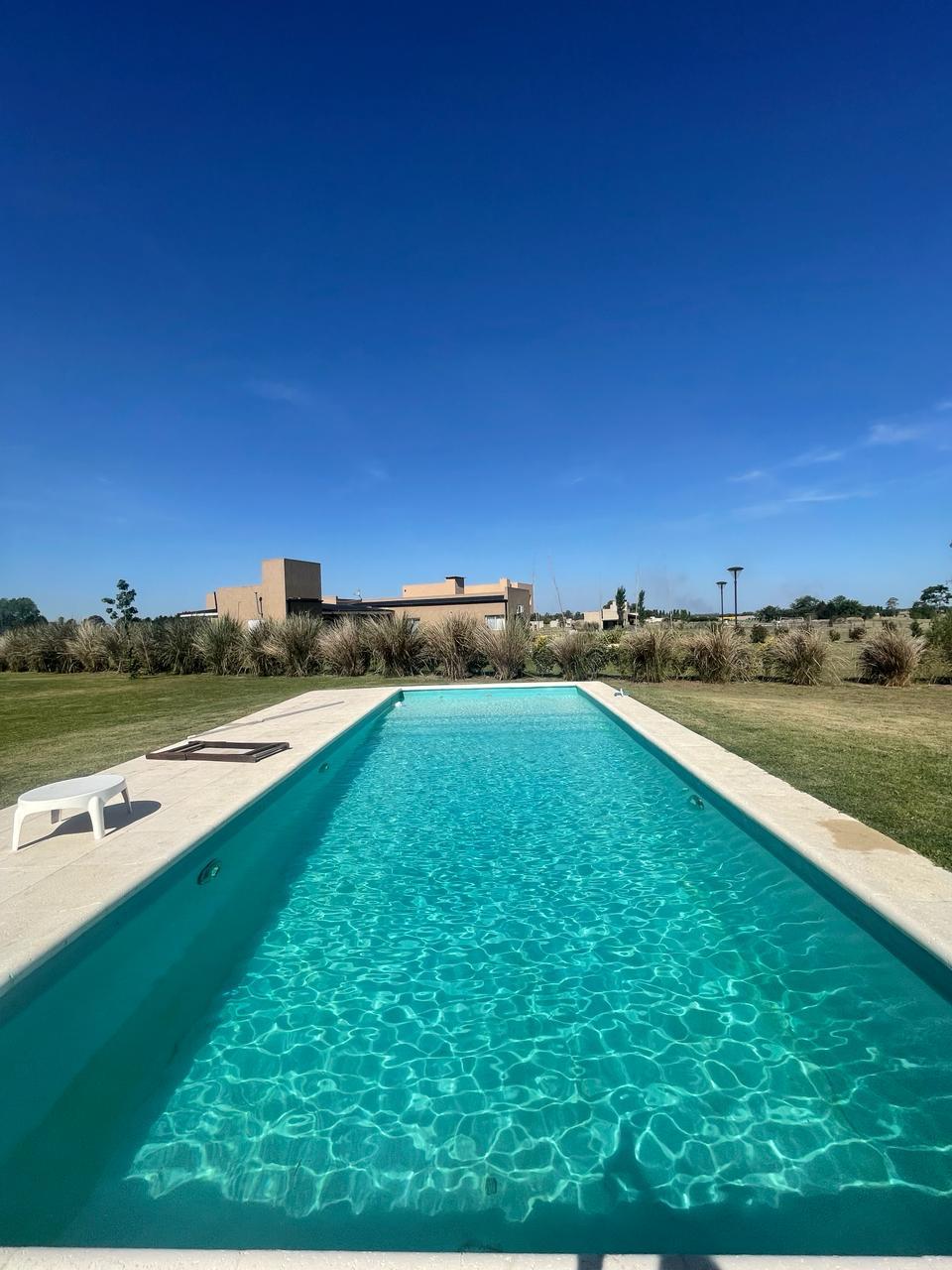 A crystal-clear swimming pool stretches across a spacious outdoor area, surrounded by lush greenery. Sunlight reflects off the water's surface, highlighting its inviting turquoise hue. A white lounge chair is positioned nearby, enhancing the space's appeal for relaxation and leisure.