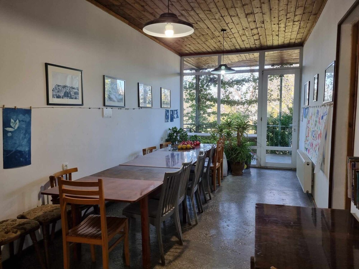 A bright dining area showcases a long table surrounded by wooden and metal chairs. Large windows offer views of the greenery outside, while framed artwork adorns the walls. Indoor plants add a touch of nature, enhancing the welcoming environment.