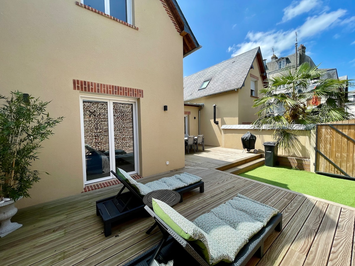 A spacious outdoor terrace is depicted, featuring two lounge chairs positioned on wooden decking. A small table sits between the chairs. A green patch of grass adds a natural touch, while a residential building can be seen in the background under a clear blue sky.