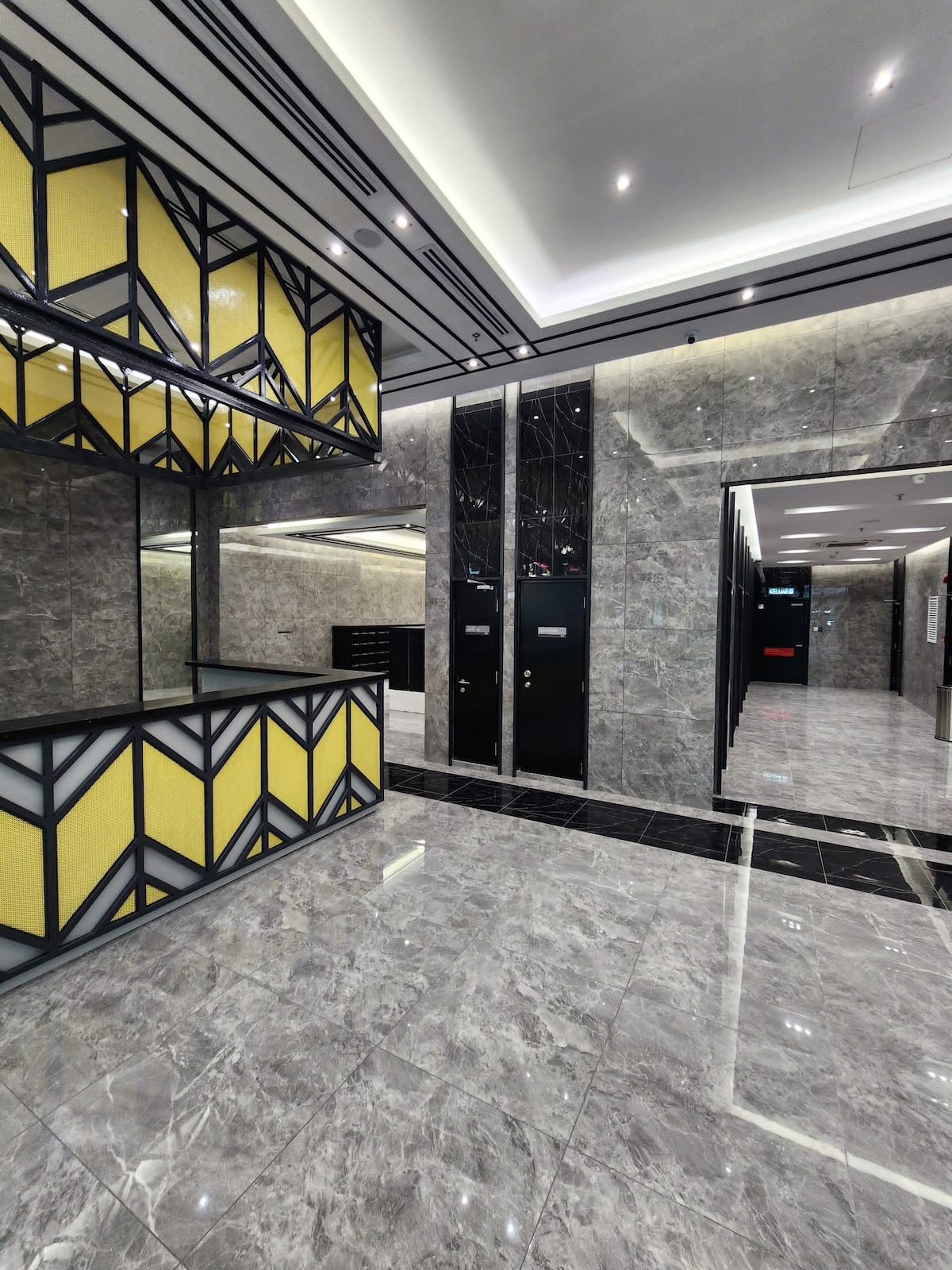 The lobby features a sleek design with polished marble floors and a striking geometric reception counter in black and yellow. Large mirrors enhance the space, while elegant lighting fixtures illuminate the area, creating a modern atmosphere for guests.