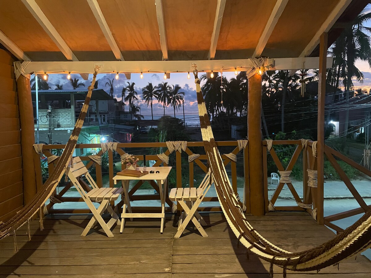 A cozy balcony is presented with two hammocks and a small table surrounded by four chairs. Soft lighting from string lights enhances the tranquil atmosphere, while views of palm trees and a street can be observed in the background.