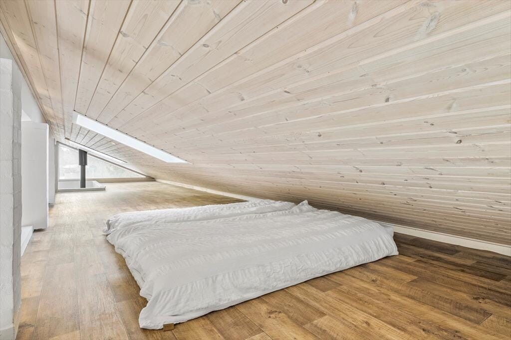 A light and airy loft space features a low ceiling lined with wooden planks and large skylights allowing natural light to illuminate the area. A spacious, unmade bedding arrangement rests comfortably on the hardwood floor, enhancing the cozy atmosphere.