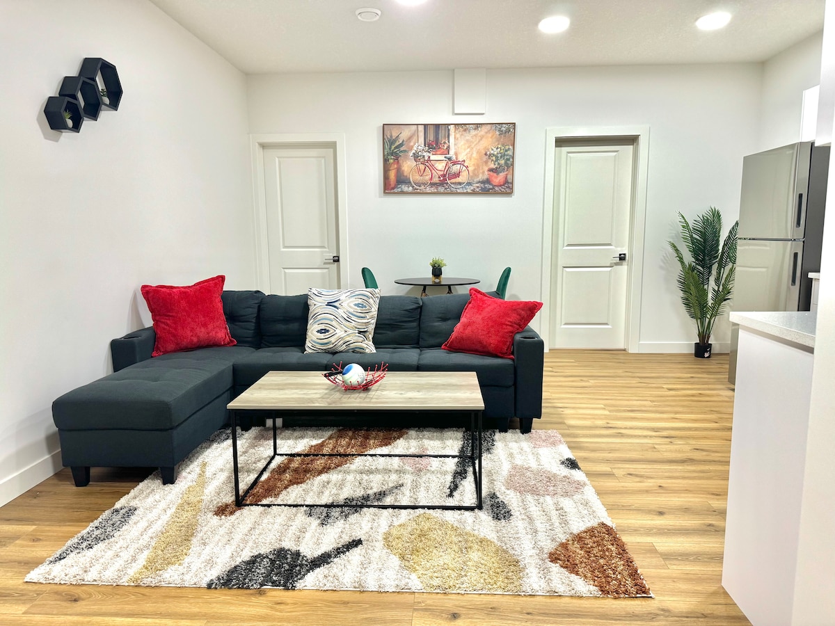 A cozy living area features a dark sectional sofa adorned with red and patterned cushions. A modern coffee table sits in the center, displaying decorative items. A textured rug adds warmth to the wooden floor, while artwork and indoor plants enhance the space's inviting ambiance.